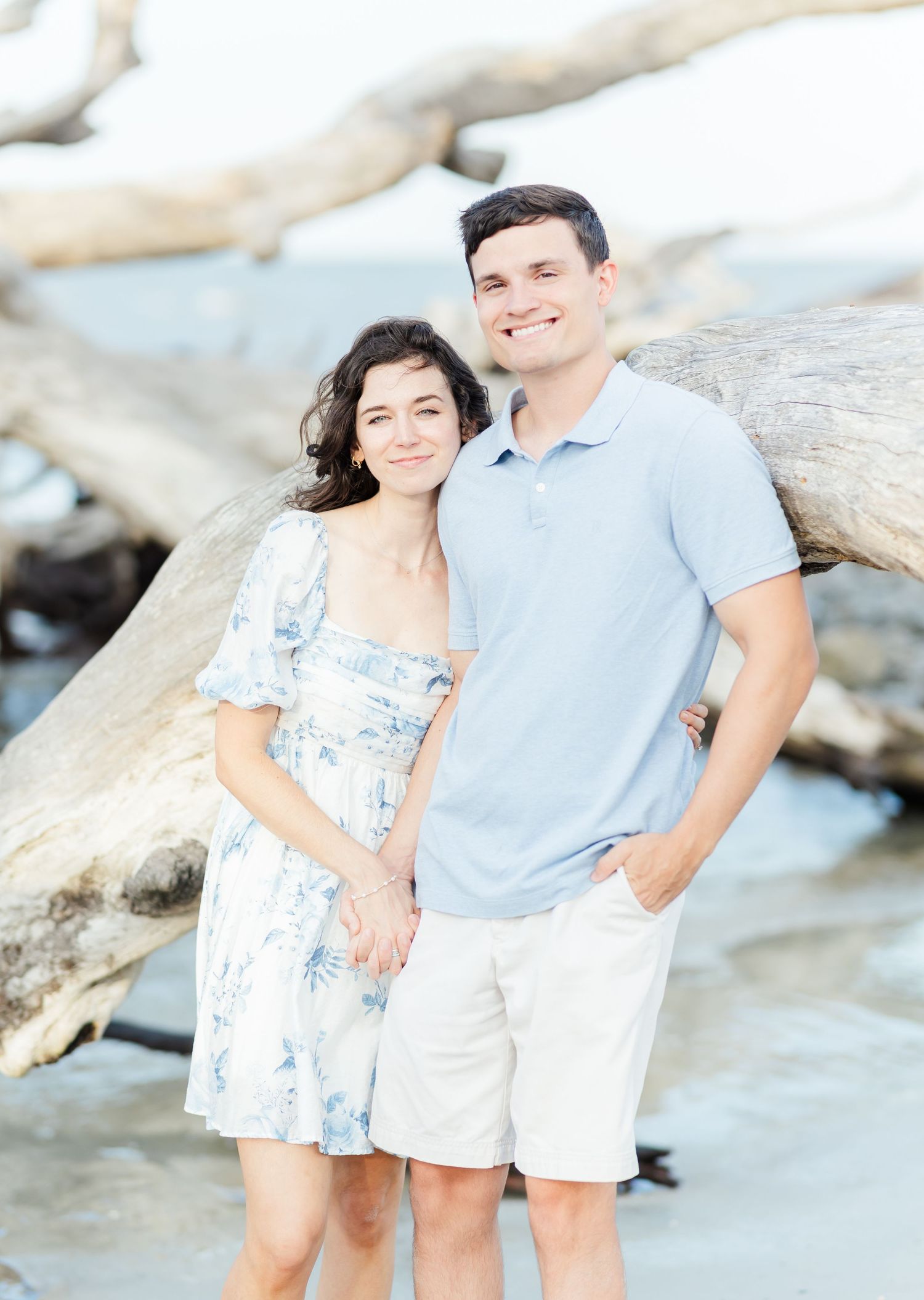 Couple in coordinated blue and white outfits pose casually against beach driftwood.