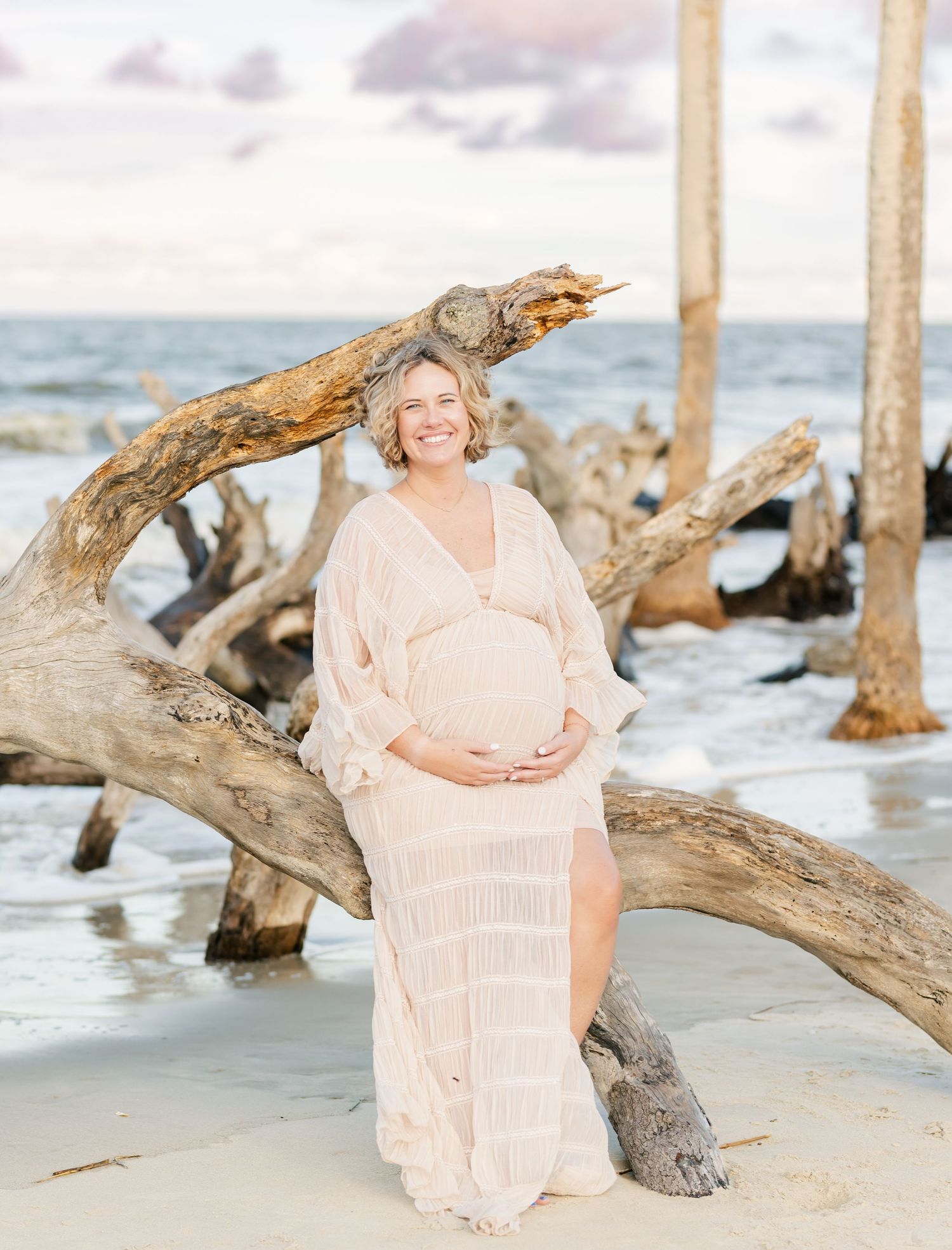 Maternity photos on a driftwood-covered beach at sunset show a person in a flowing peach dress sitting on weathered logs.