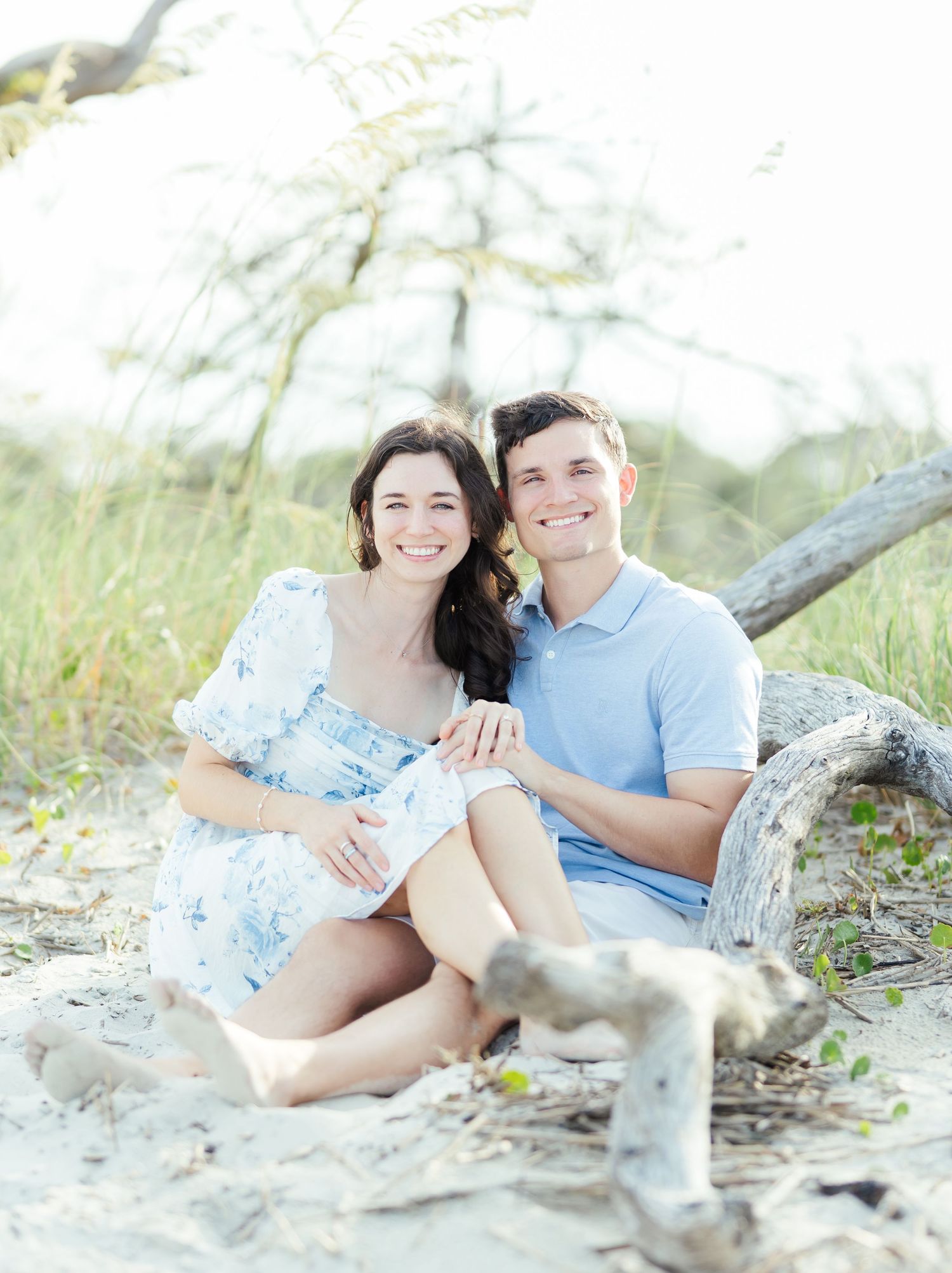 A couple sits together by driftwood on a sandy beach wearing coordinated blue and white outfits.