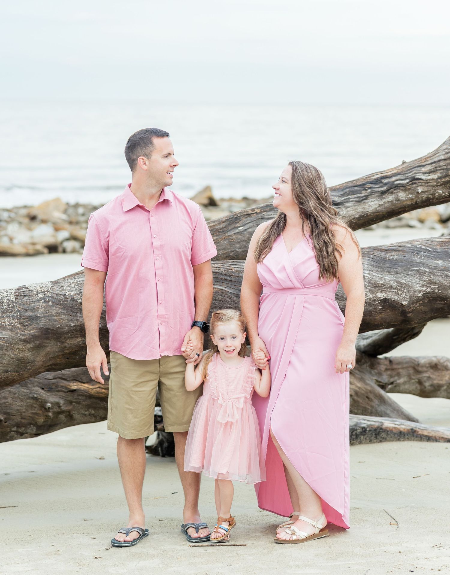 A family in coordinating pink outfits smiles together while standing on a beach with driftwood and rocks in the background.