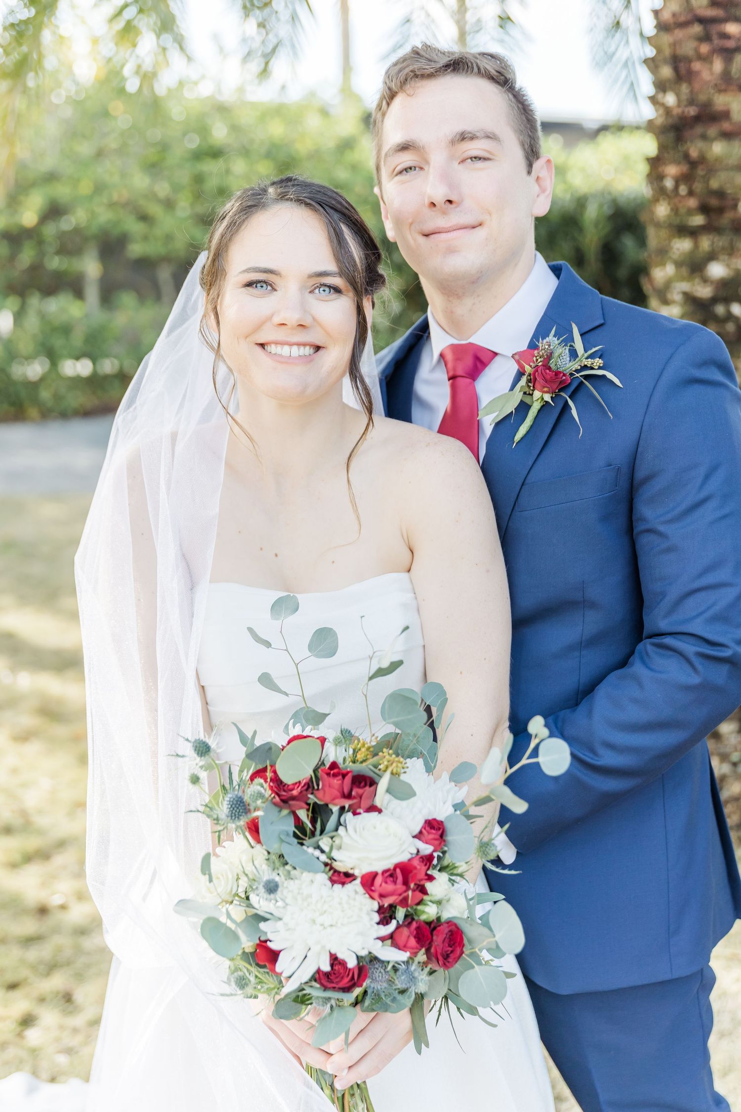 A sunny outdoor wedding portrait with bride holding red and white floral bouquet.