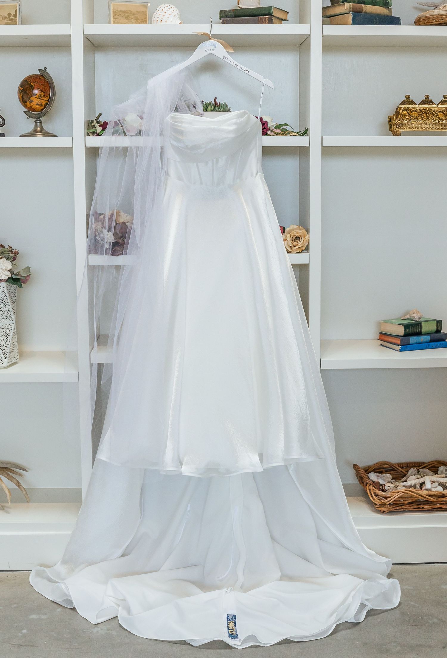 White wedding gown displayed on hanger against built-in shelves with vintage decor.