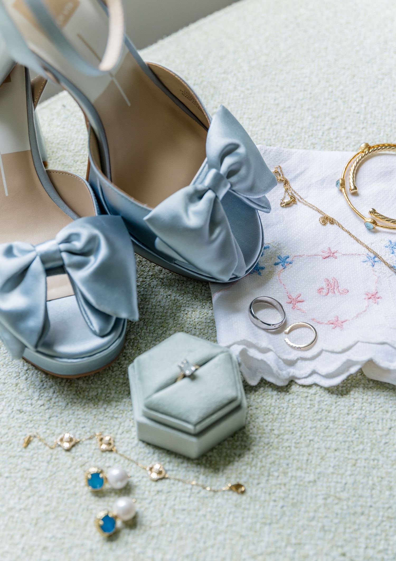 Light blue satin bow heels with wedding rings and accessories on embroidered handkerchief.