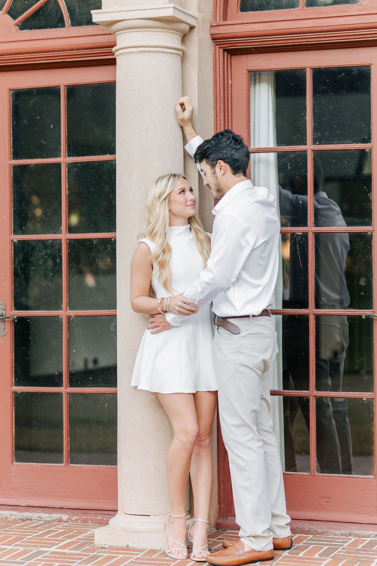 A series of romantic engagement photos showing a couple in white outfits posing against a building with coral-colored windows.