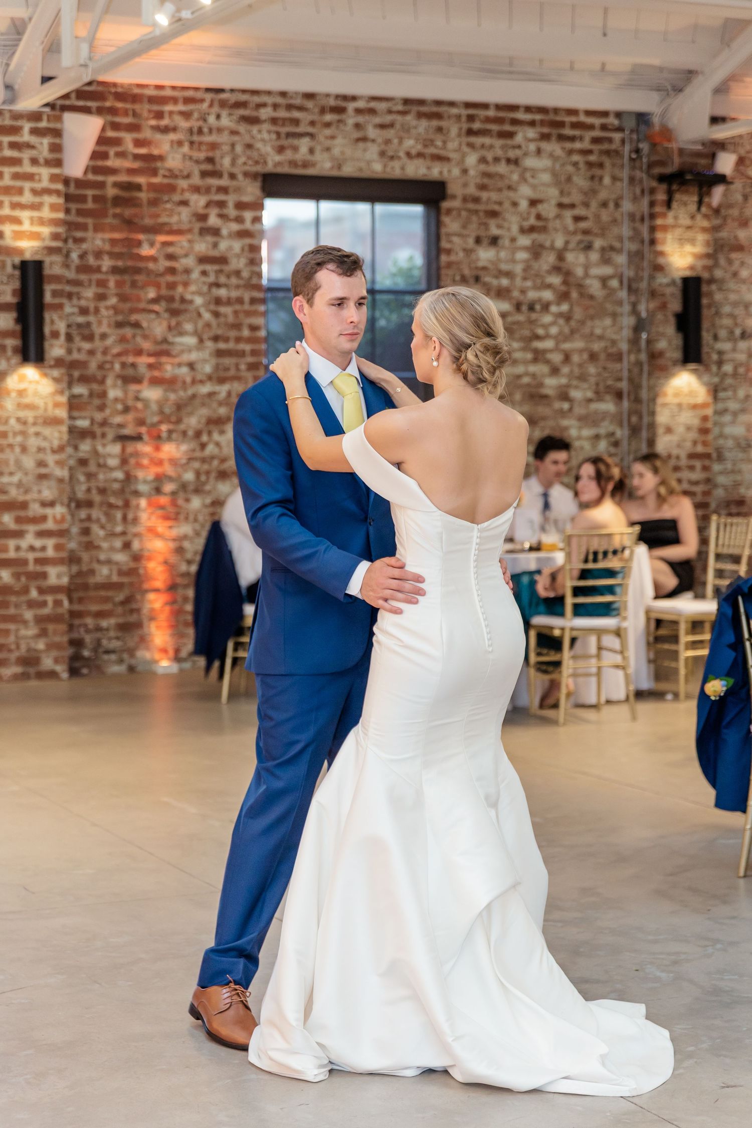 A couple shares their first dance in a rustic brick-walled venue with warm lighting and elegant decor.