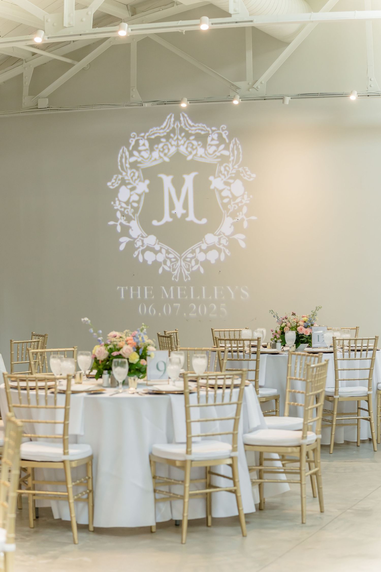 Elegant wedding reception setup with white tablecloths, gold chiavari chairs, and a monogrammed 'M' projection on the wall.
