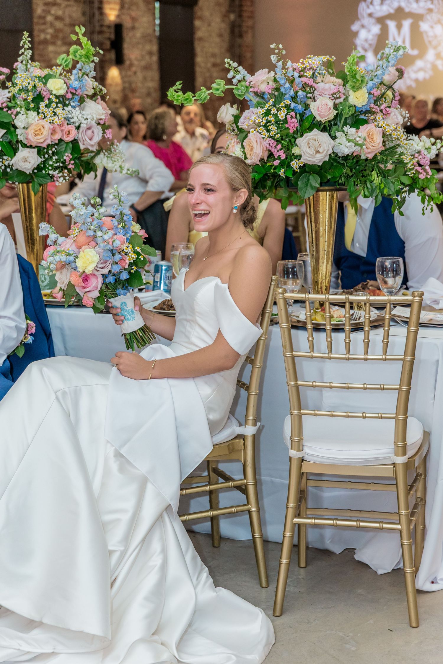 Wedding party in white dresses and gold chairs enjoying a celebration with floral centerpieces.