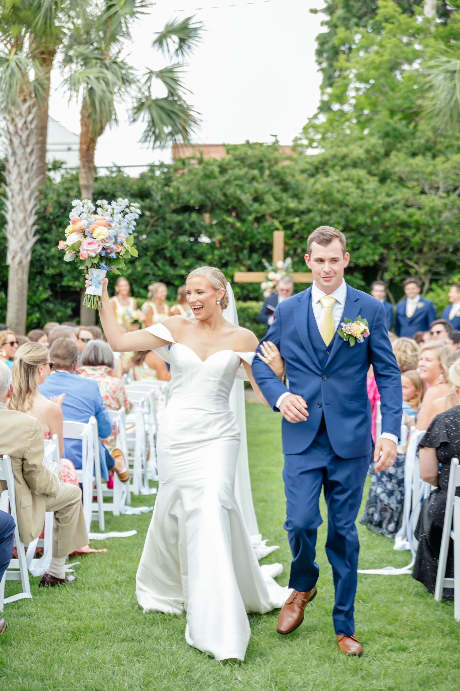 Wedding guests celebrating and cheering during an outdoor ceremony with palm trees in the background.