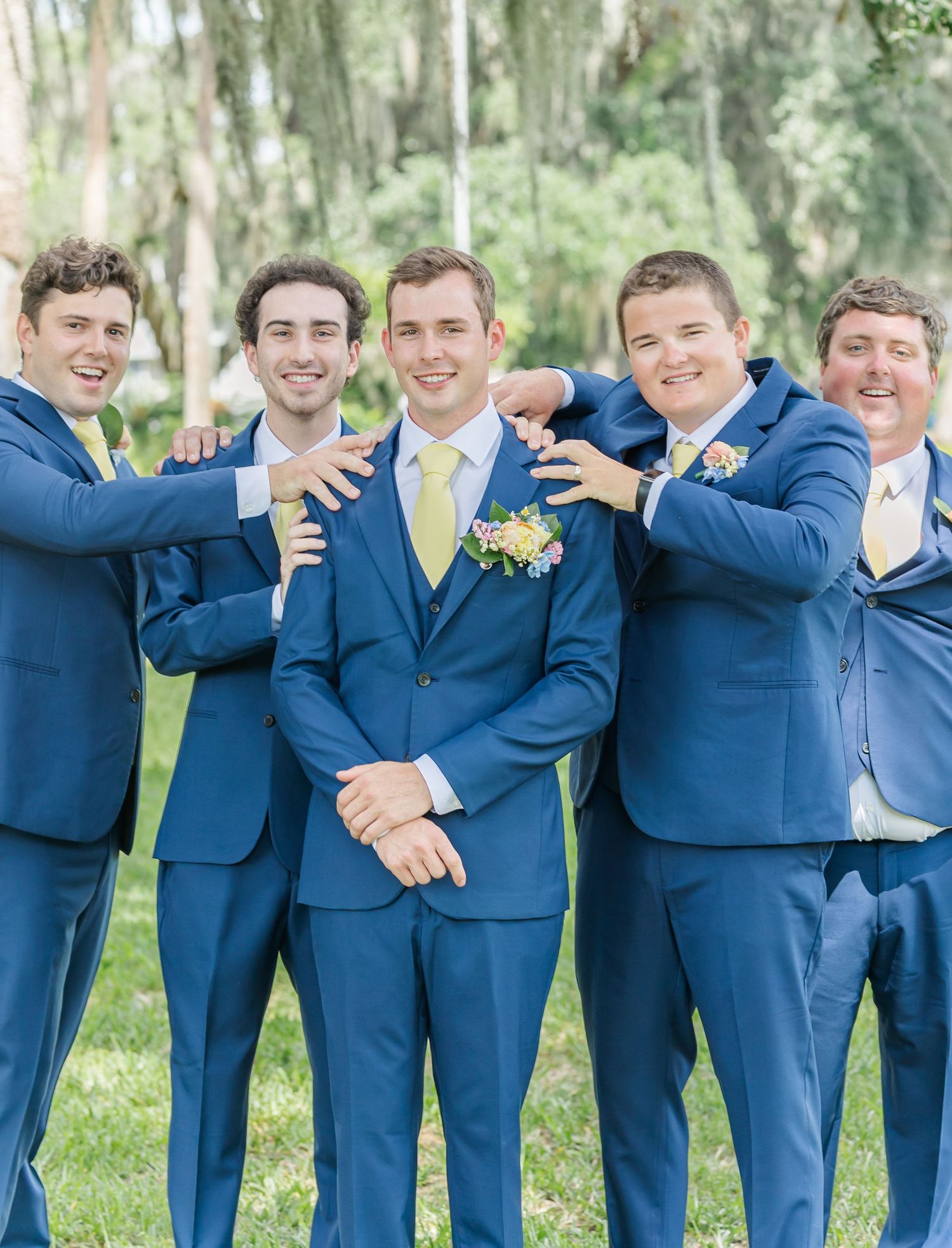Groomsmen in navy blue suits with yellow ties pose playfully together at outdoor wedding venue.