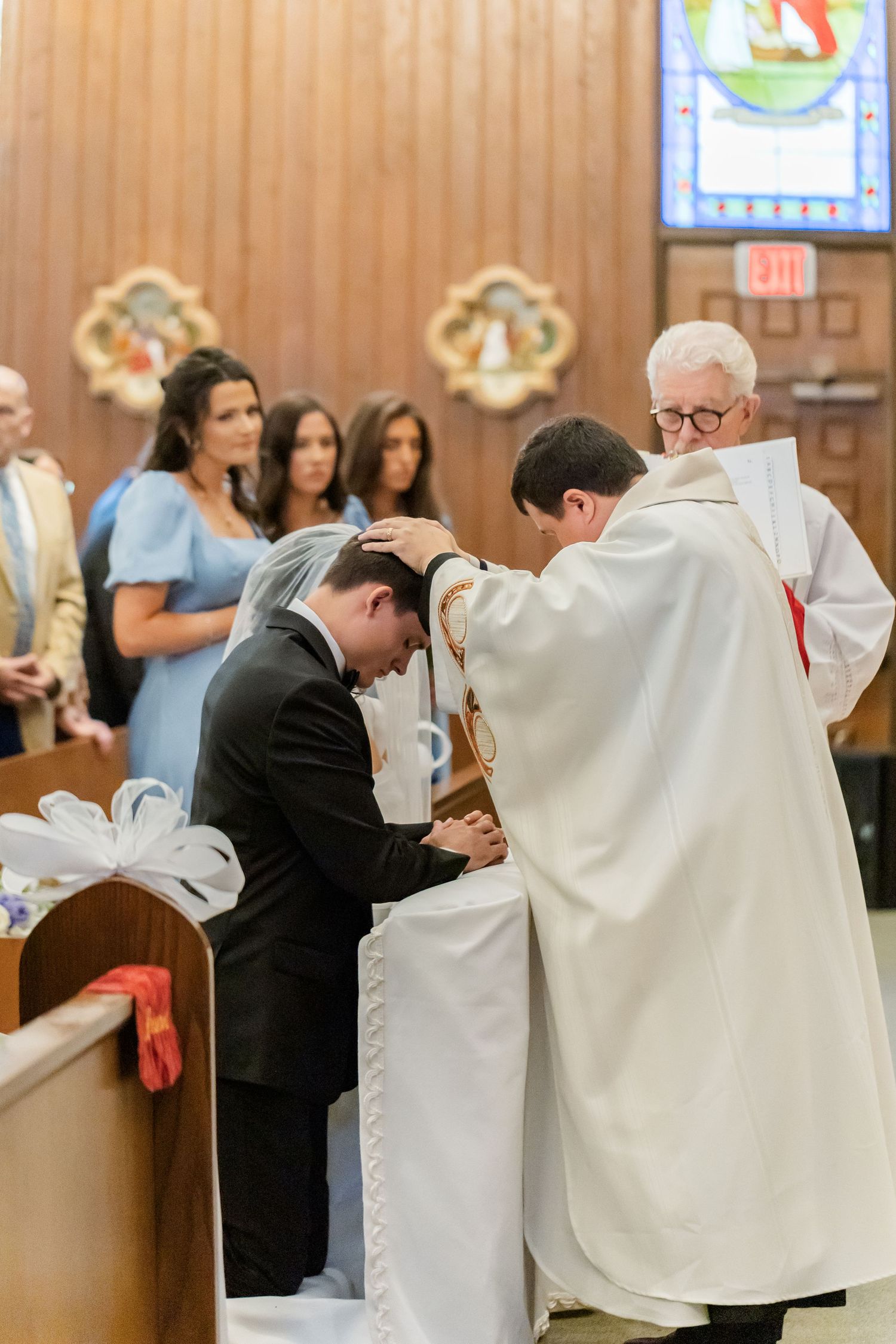 Religious ceremony with participants in white robes and dark suits kneeling in church.