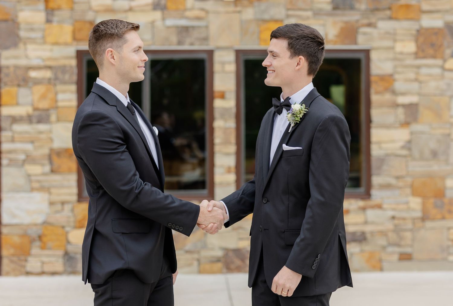 Two men in formal black tuxedos stand together against a stone wall exterior.