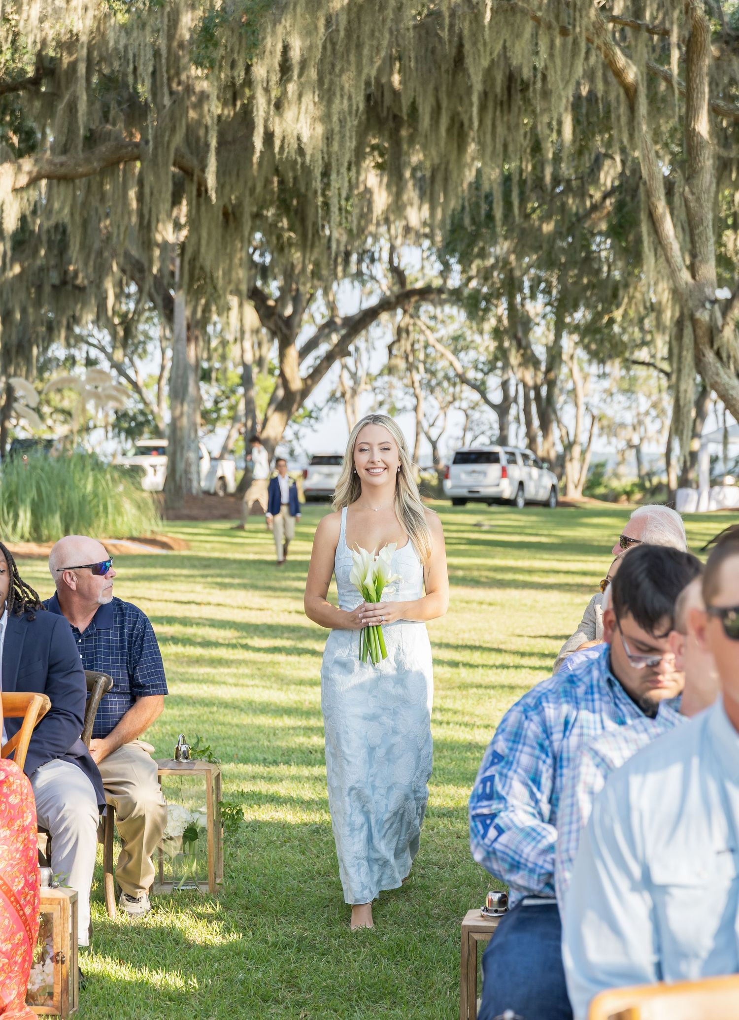 Wedding guests gather under Spanish moss-draped trees at an outdoor ceremony in a park-like setting.