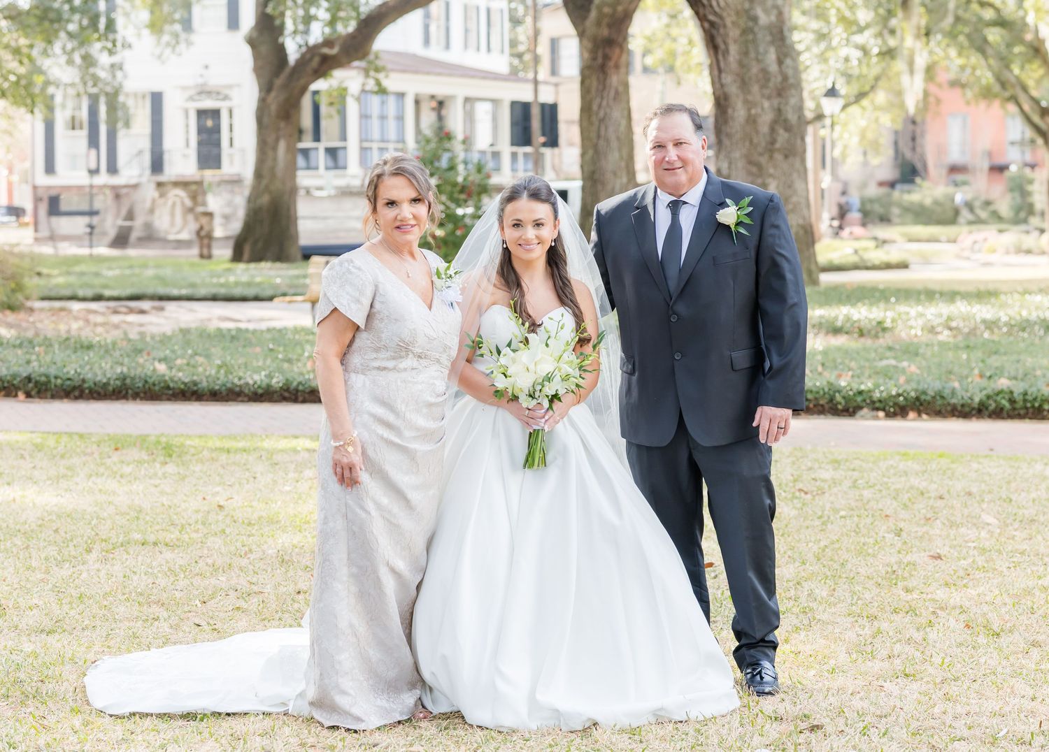 Wedding party members stand in formal poses on grassy lawn with trees and buildings in background.
