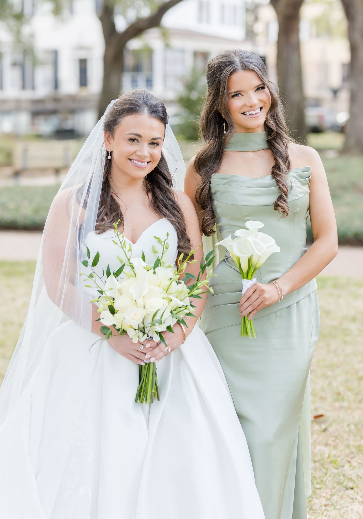 Bride and bridesmaid in sage green dress hold white floral bouquets outdoors.