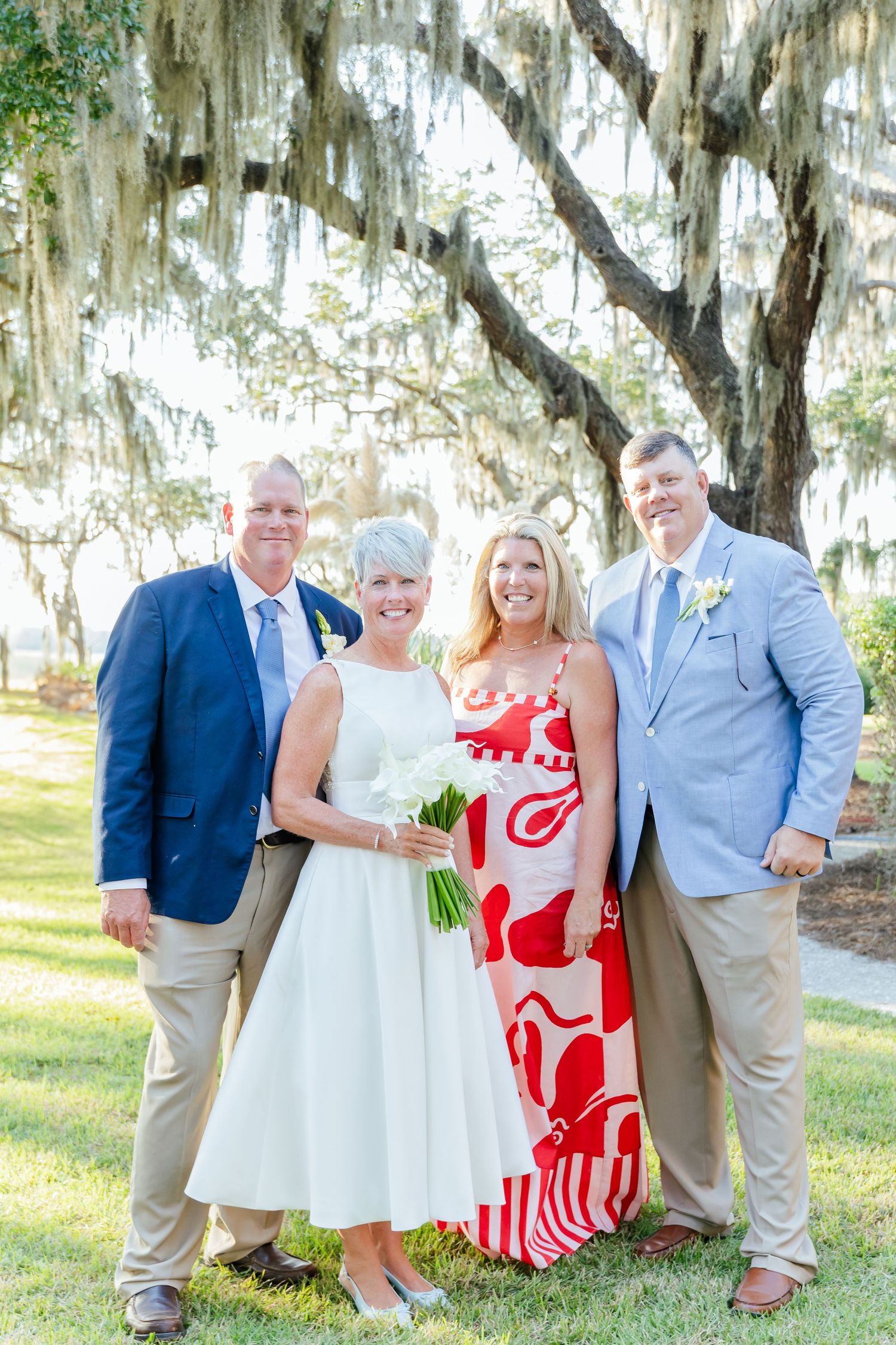 A wedding party poses outdoors under Spanish moss-draped trees in a natural setting with white and red attire.