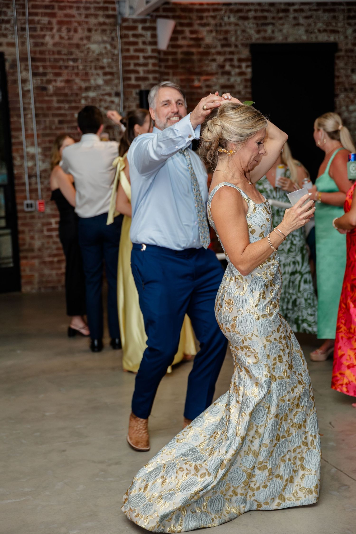Wedding guests dance energetically in a rustic brick venue with colorful dresses swirling on the dance floor.