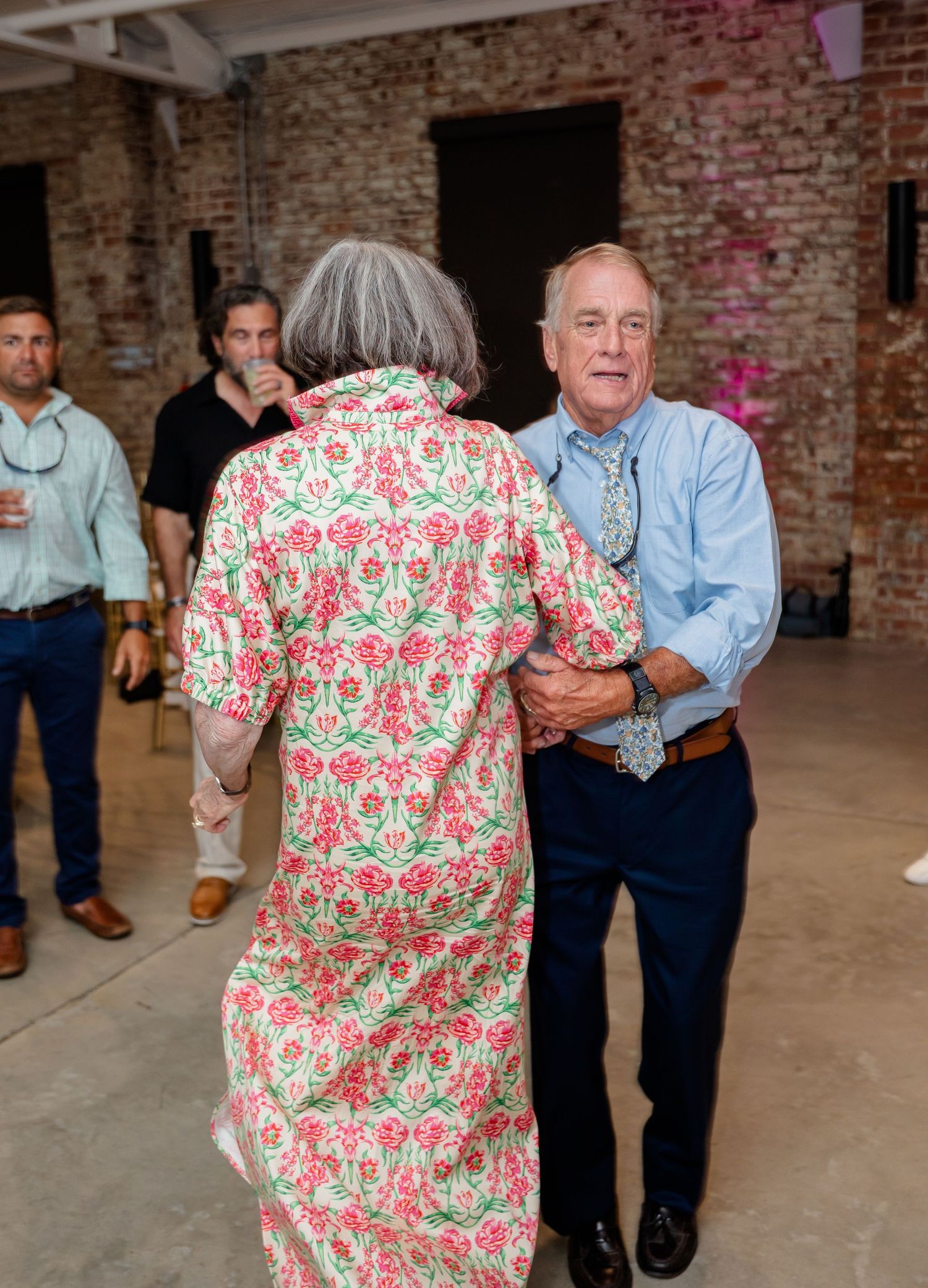 Wedding guests in floral print outfits dancing together at a rustic brick venue during the reception.