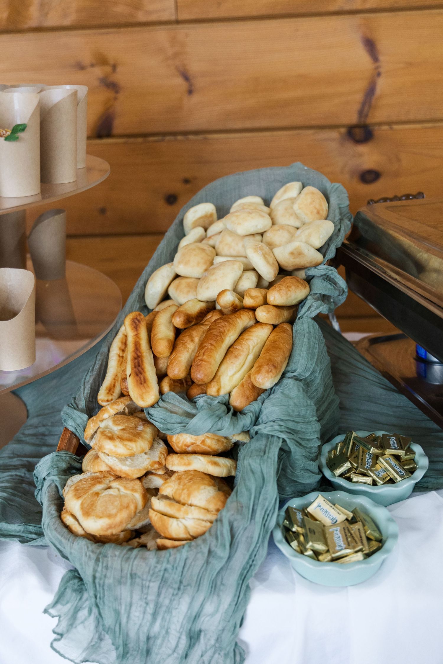 Bread rolls and pastries displayed on sage green linens with paper cups on a wooden table surface.