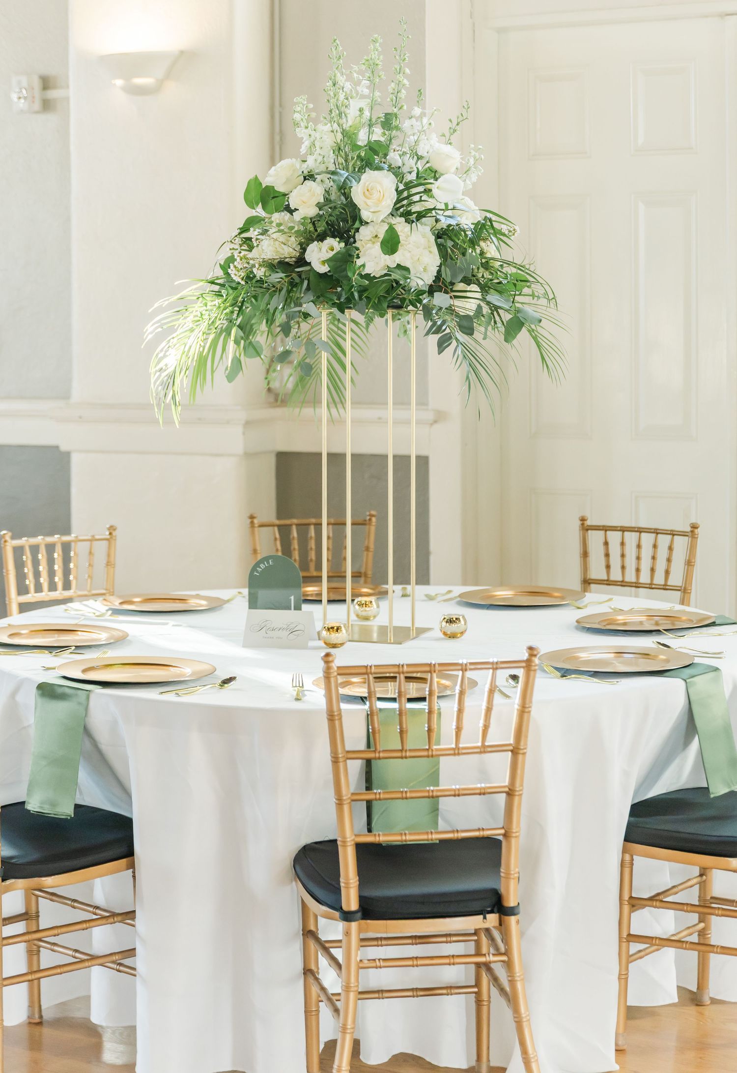 Elegant wedding reception tables with white linens, gold chiavari chairs, and lush floral centerpieces in a bright venue.