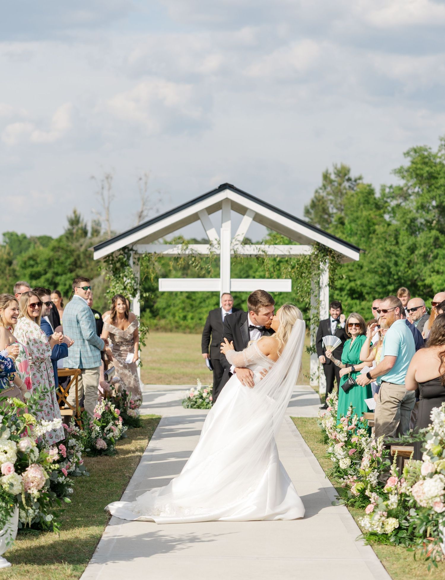 Bride and groom share first kiss at outdoor wedding ceremony with floral-lined aisle and white cross backdrop.