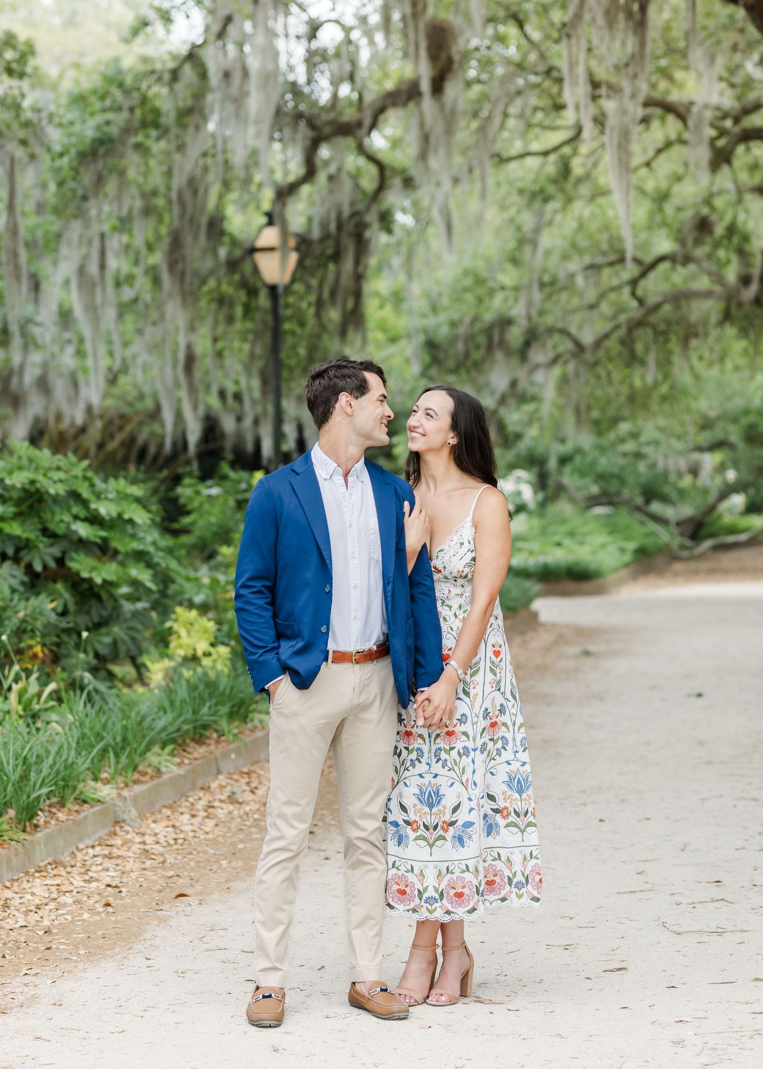 A couple smiles at each other on a tree-lined path, man in blue blazer, woman in floral dress.