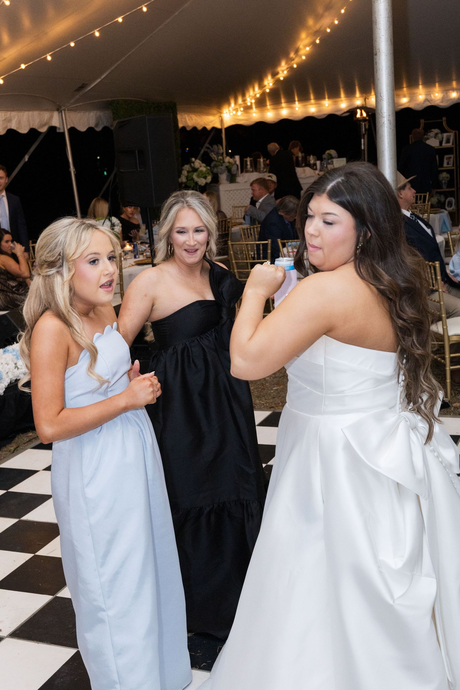 Bride dances and laughs with guests in blue and black dresses at a tented wedding reception.