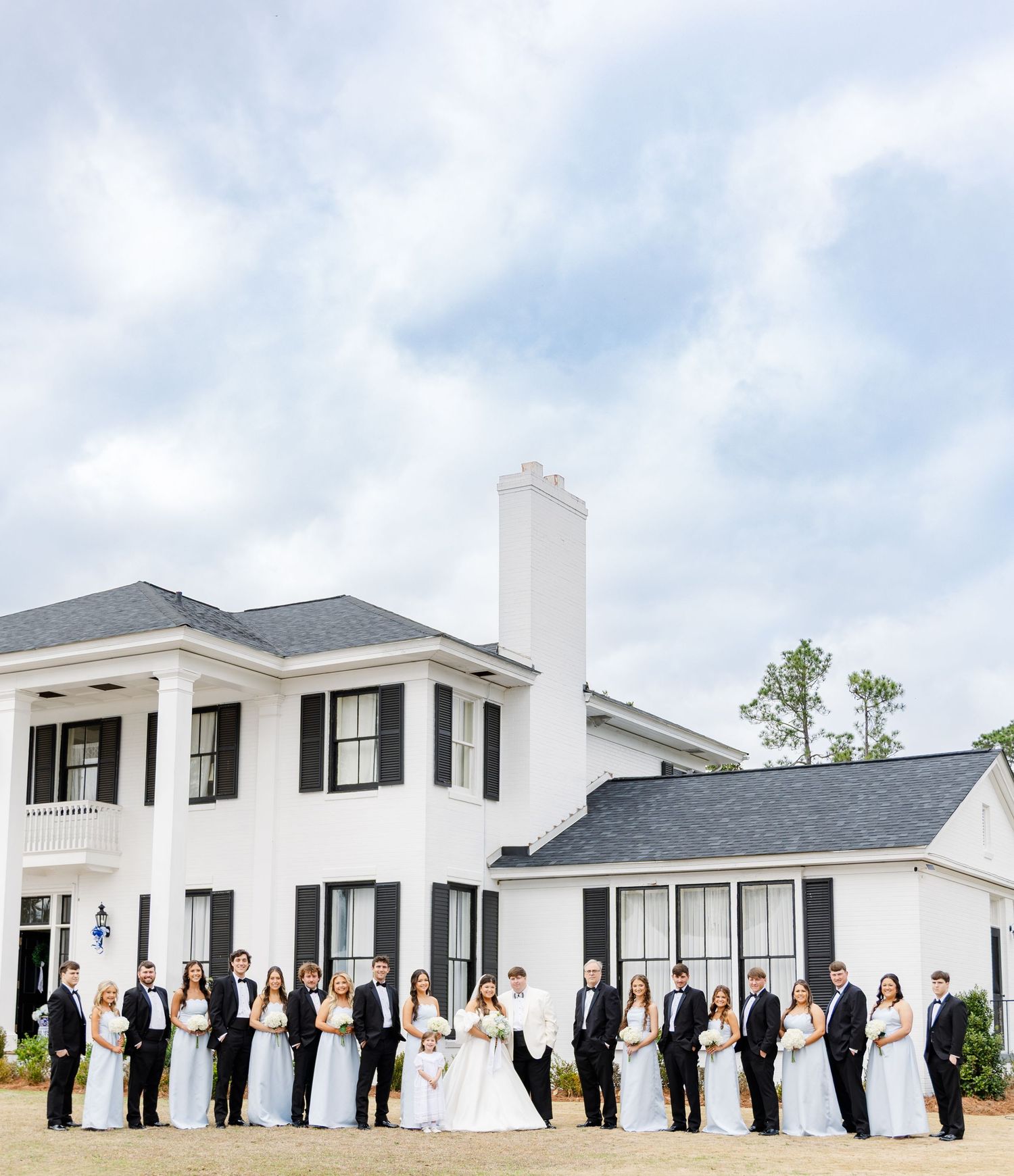 Large wedding party poses in front of grand white Southern mansion with black shutters under dramatic cloudy sky.