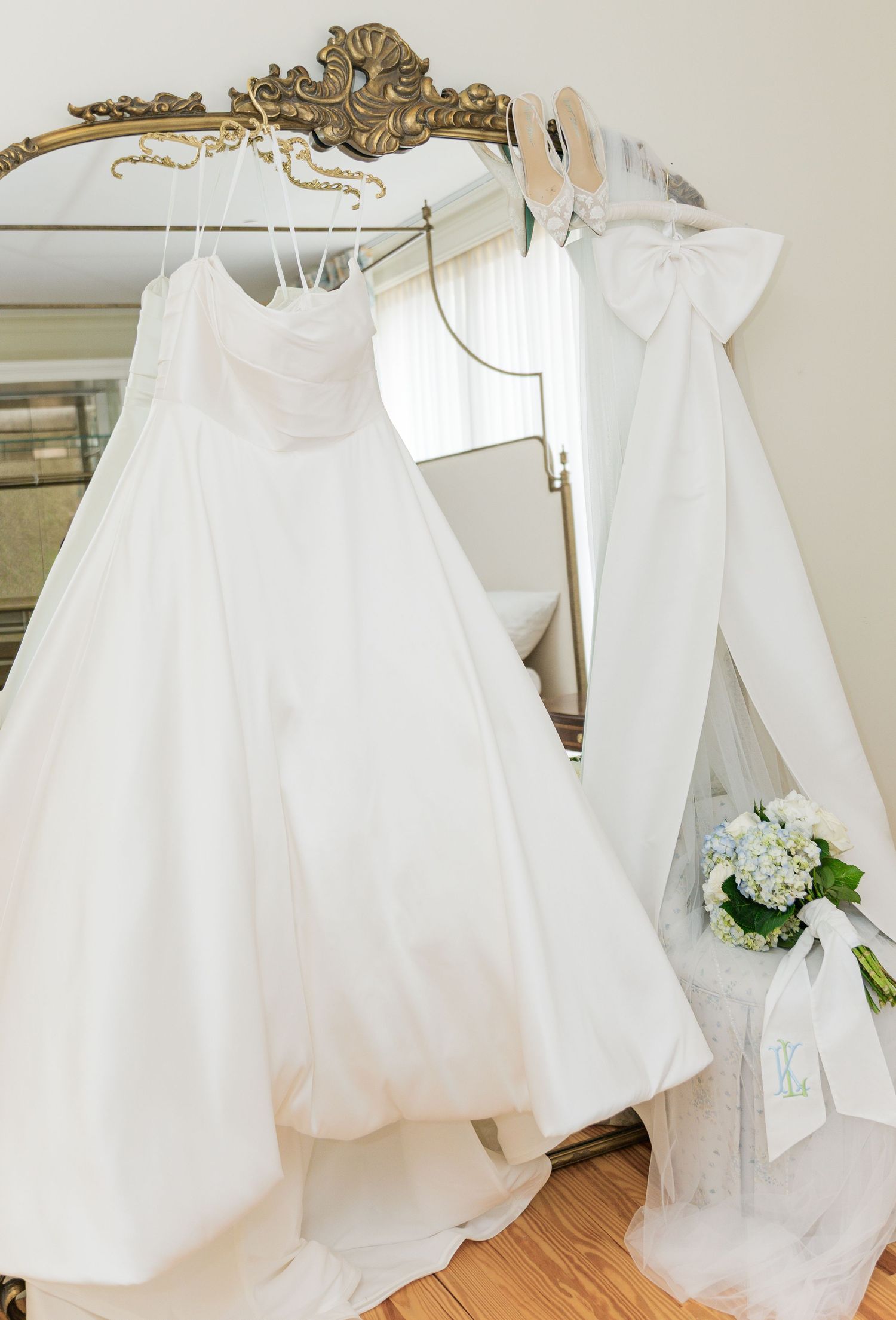 White wedding gown and flower girl dress hanging on ornate gold mirror with bridal bouquet nearby.