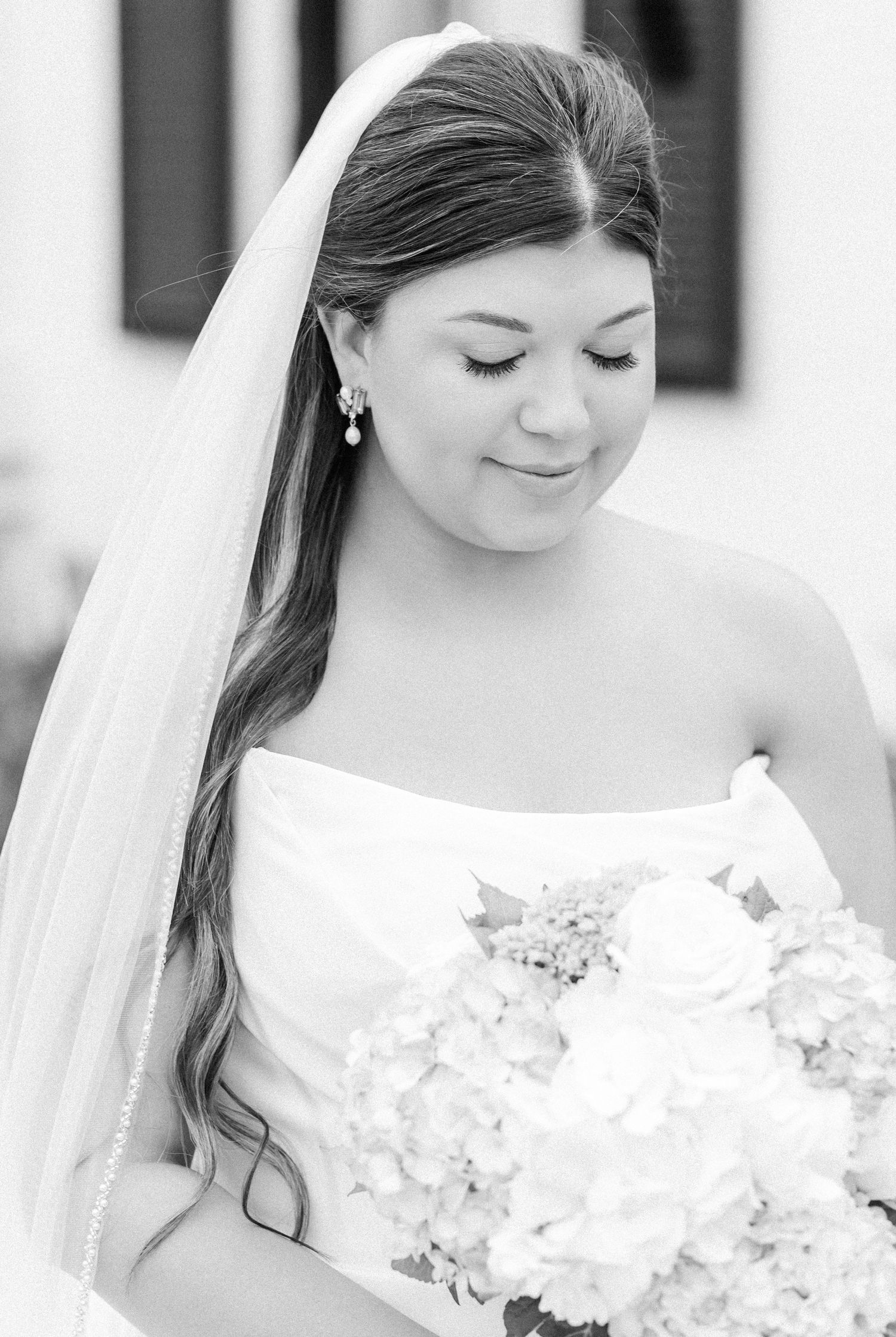 Black and white close-up portrait of bride in strapless gown with veil and pearl earrings holding bouquet.