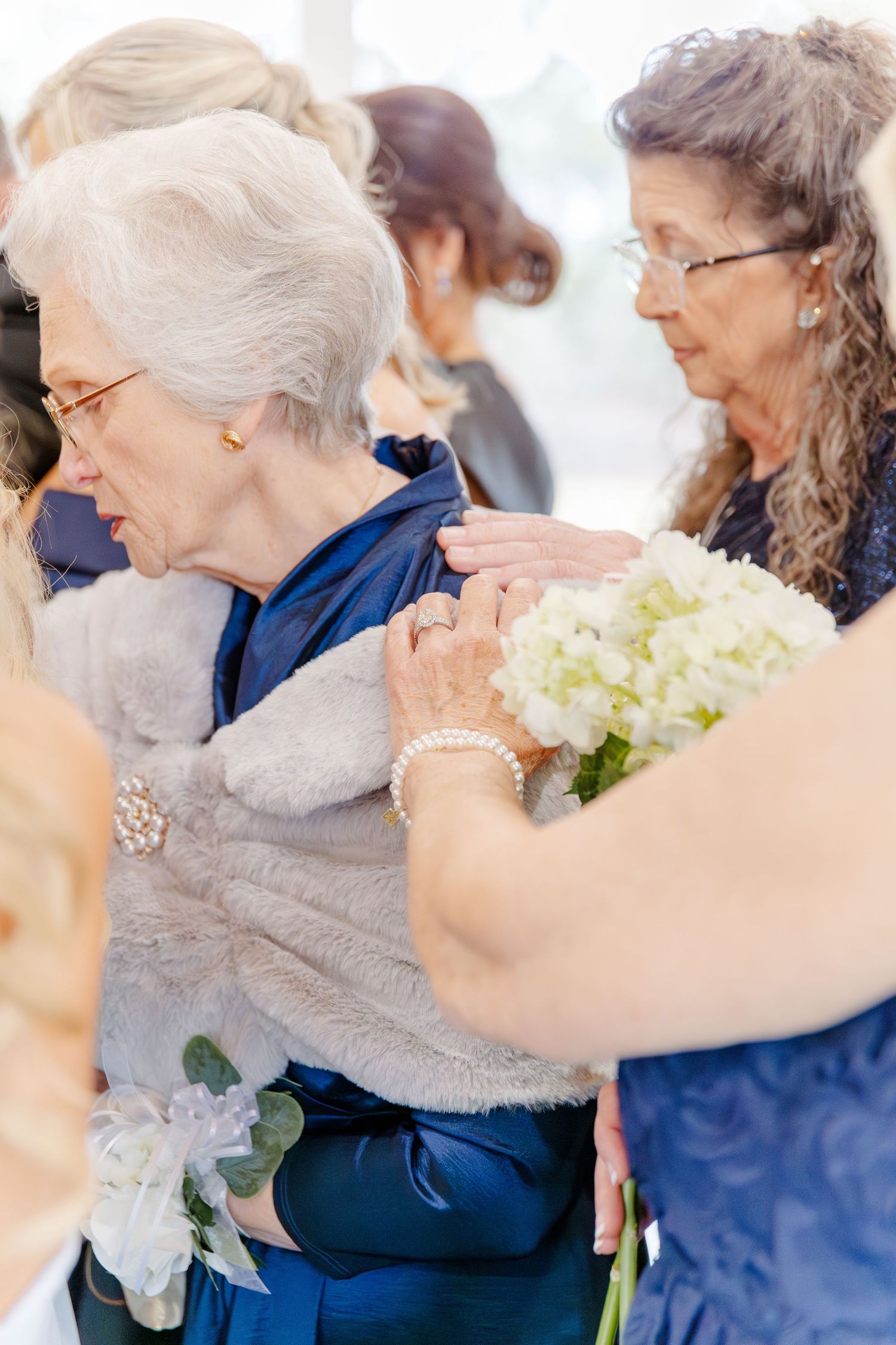 Two elderly women in blue and gray attire share an emotional moment holding white hydrangea bouquets.