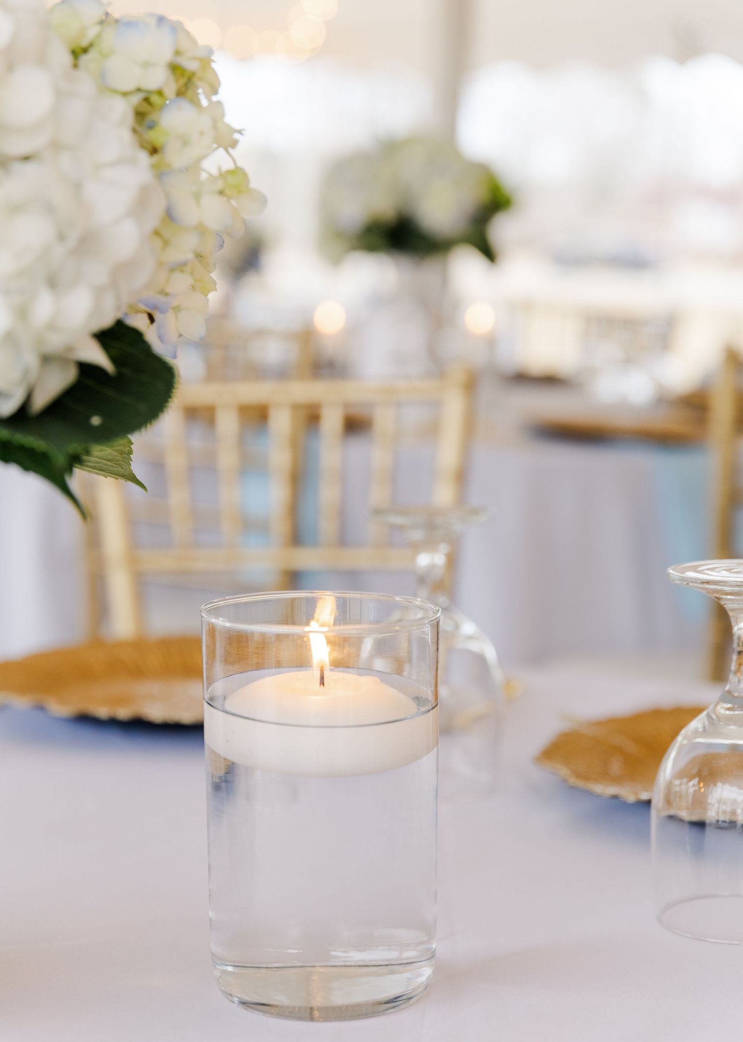 Floating candle in glass vase centerpiece with white hydrangeas and gold charger plates at reception.