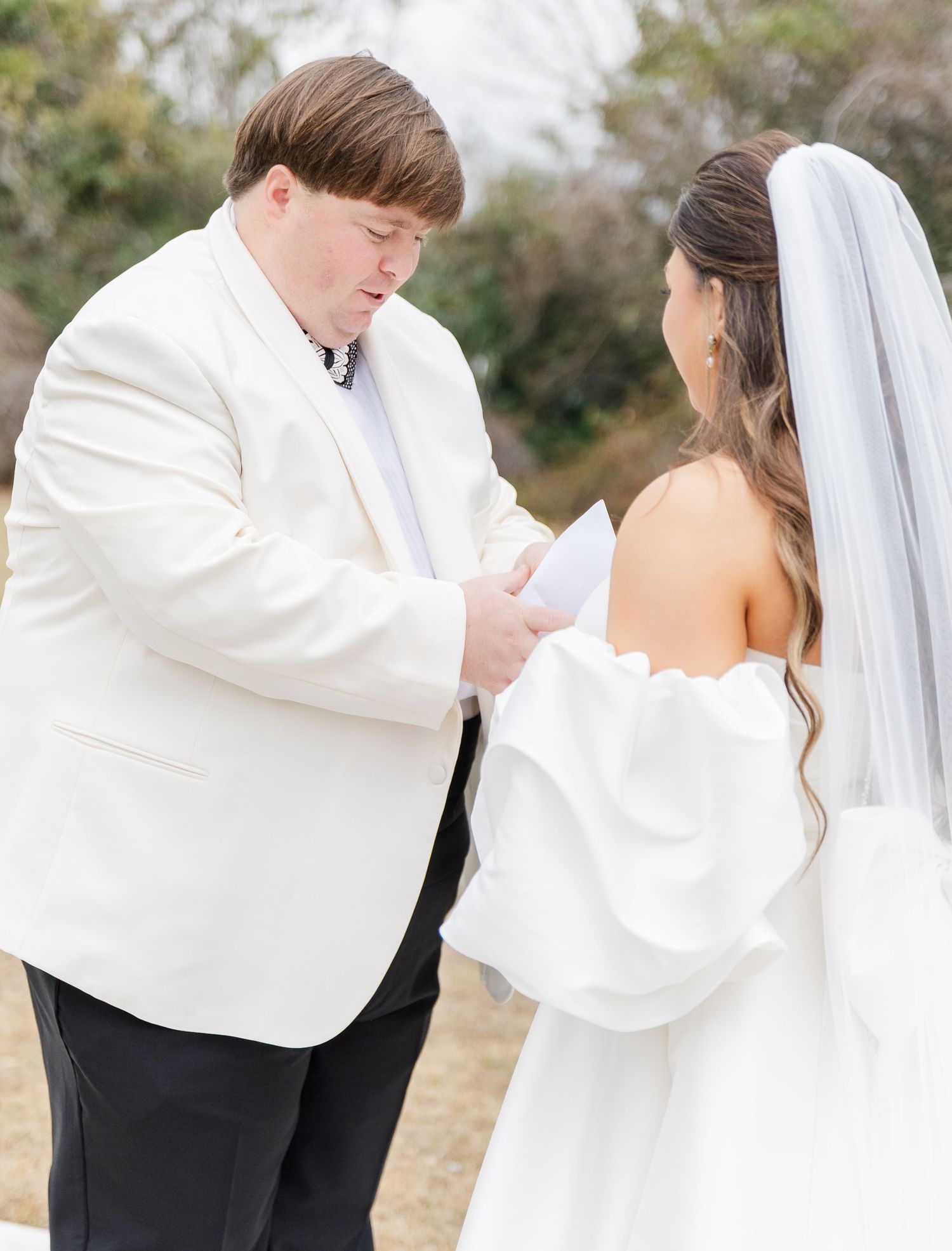 Groom in white tuxedo jacket reading vows to bride in strapless gown with veil during outdoor wedding ceremony.