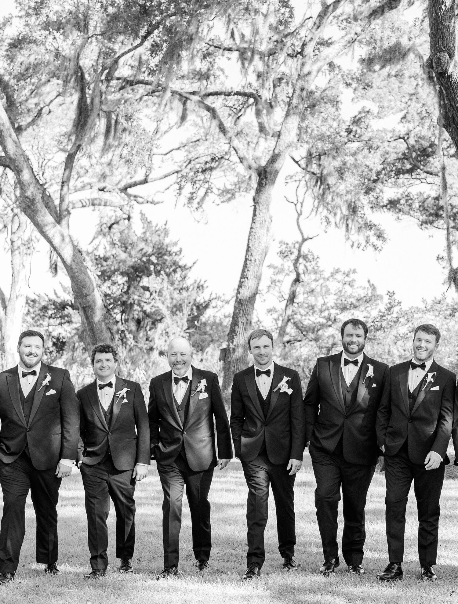 Black and white photo of groom and groomsmen in tuxedos with bow ties standing outdoors under large oak trees.