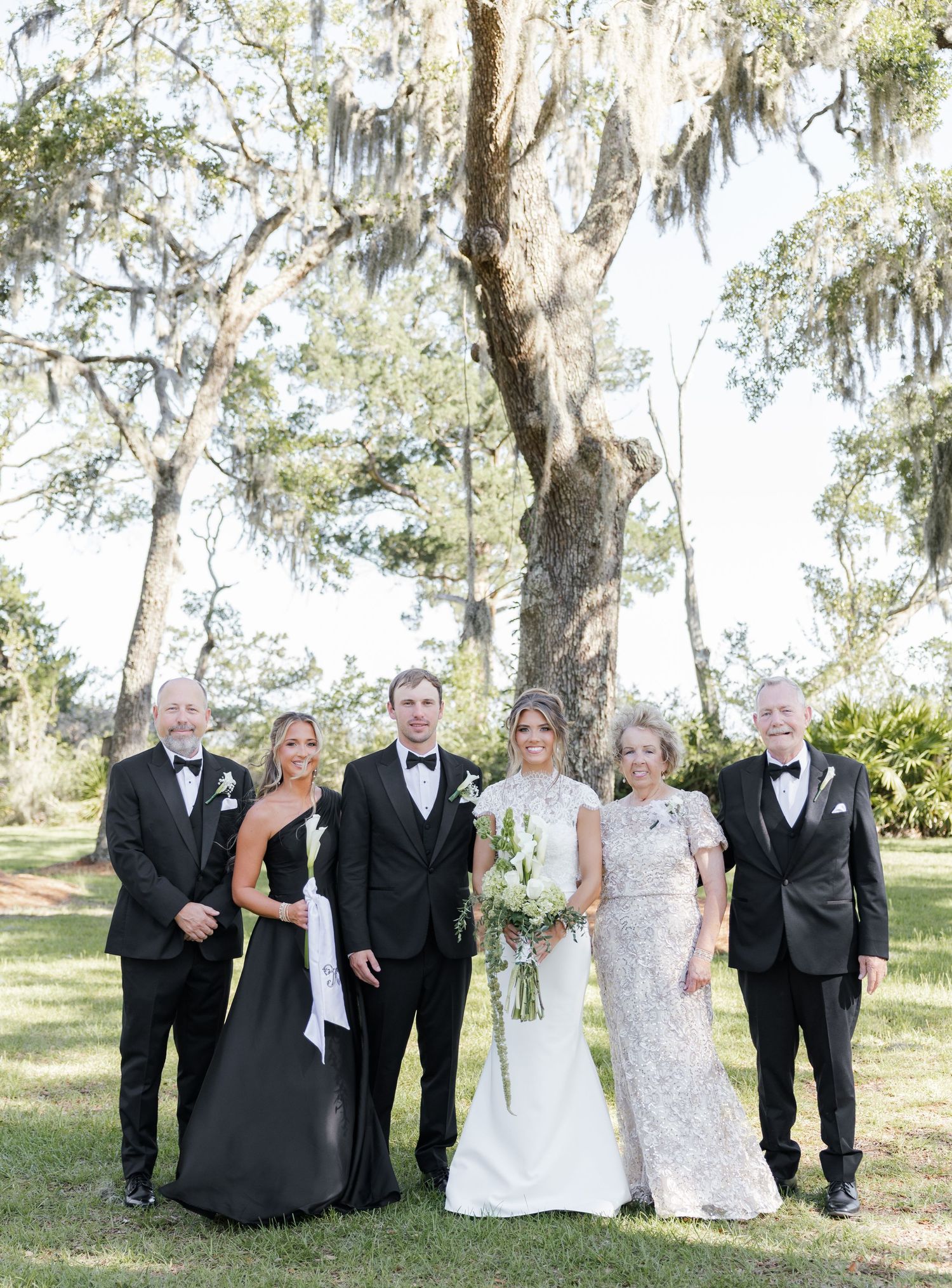 Wedding party of six poses outdoors under Spanish moss-draped oak trees, bride in white gown holding bouquet.