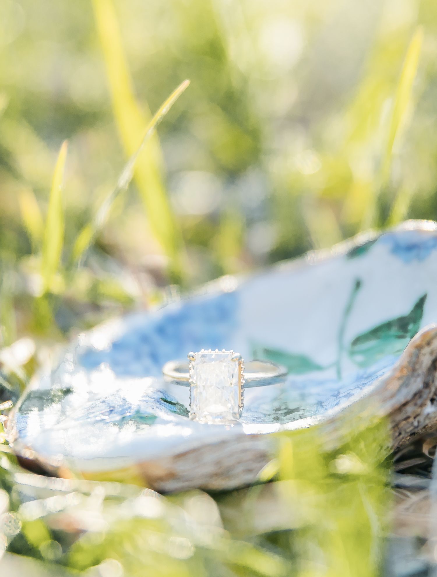 Close-up of a radiant-cut diamond engagement ring nestled on a blue surface amid grass.