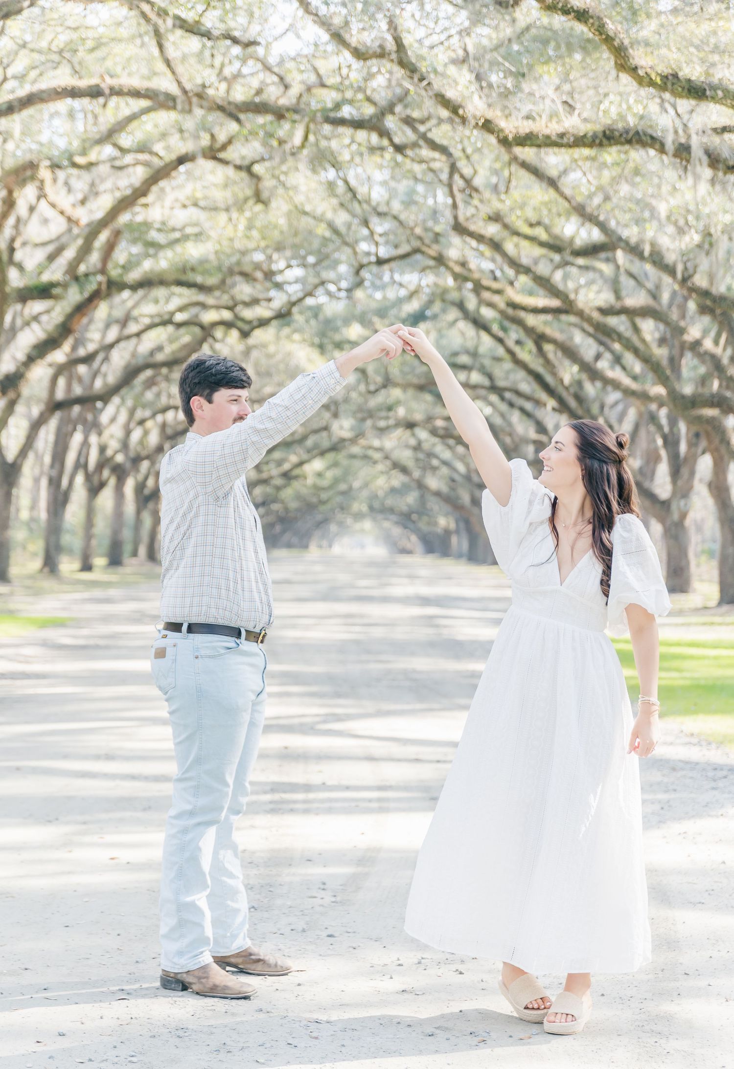 A couple reach up to touch hands while dancing on a tree-lined path.