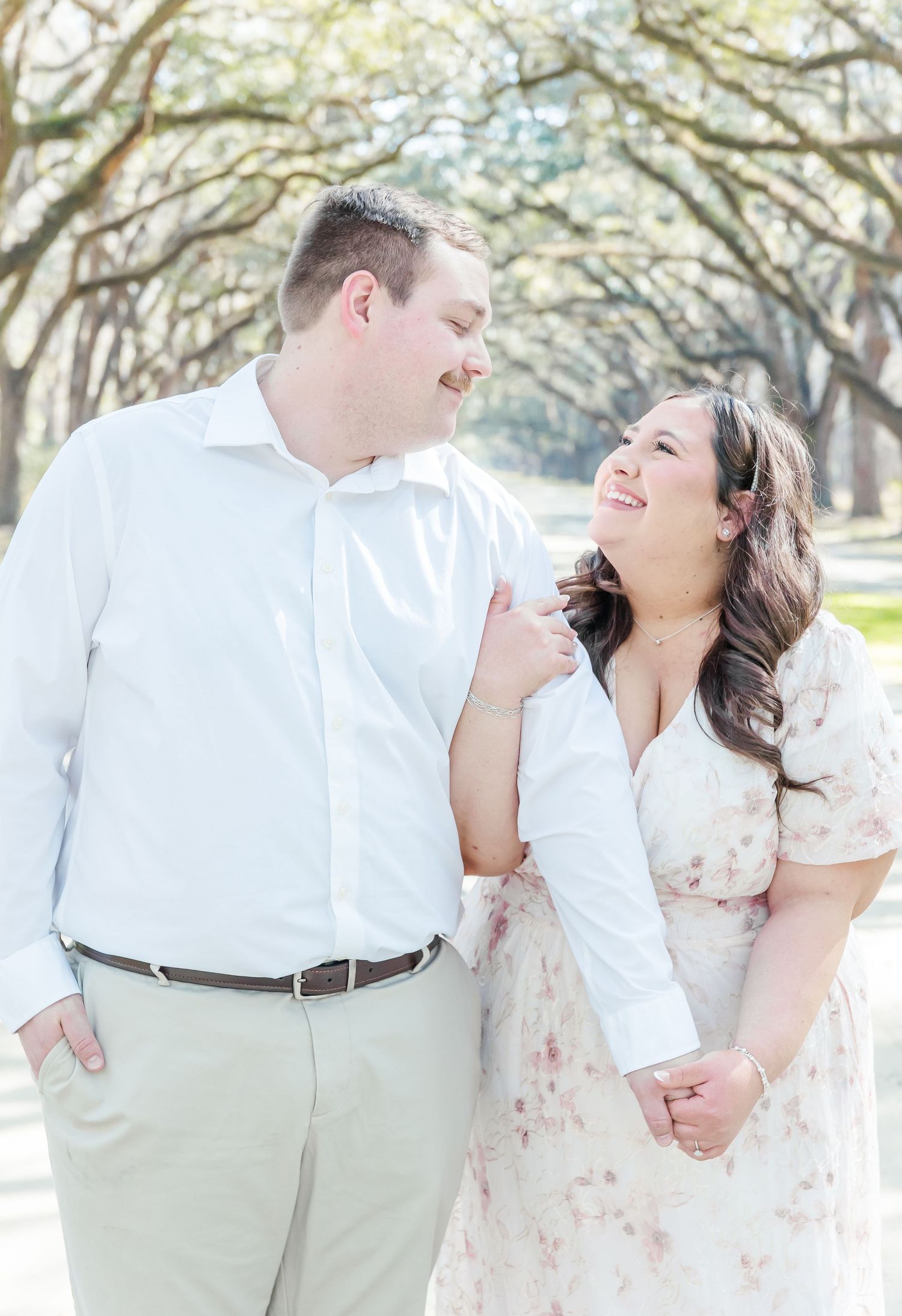 Couple laughing together during engagement photos beneath oak trees.