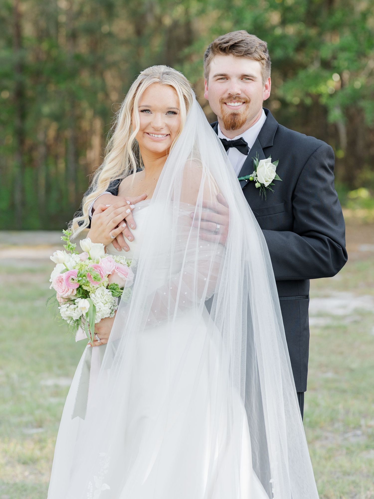 A smiling bride in a white gown and veil holds a pink bouquet beside a groom in a black tuxedo outdoors.