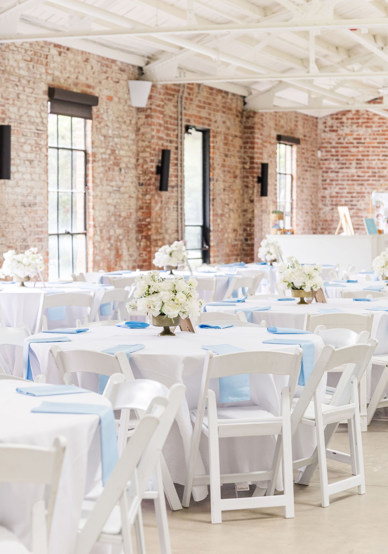 Elegant wedding reception with white chairs, blue accents, floral centerpieces, and exposed brick walls.