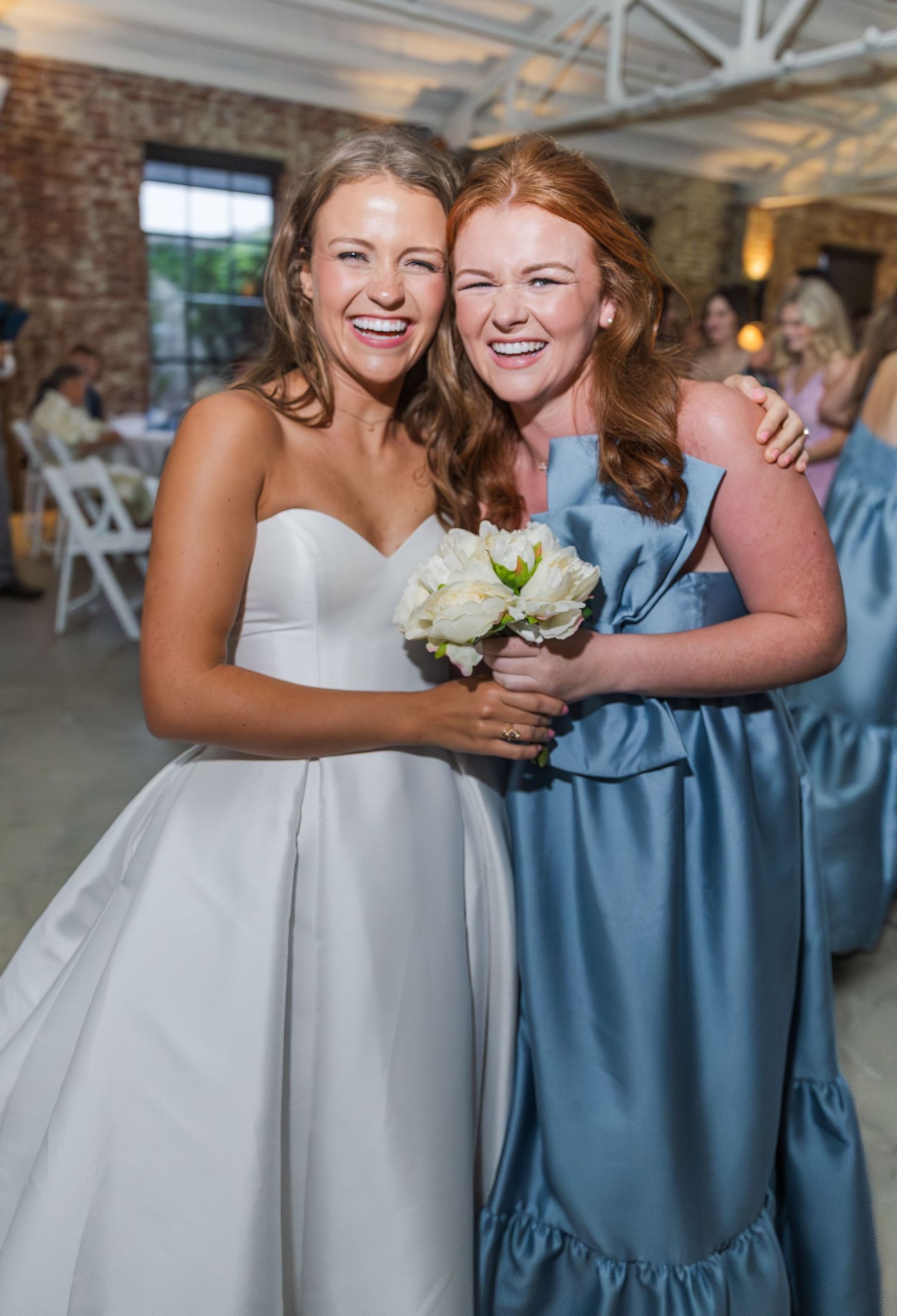 Smiling bride in white strapless gown poses with bridesmaid in blue dress holding white bouquet.