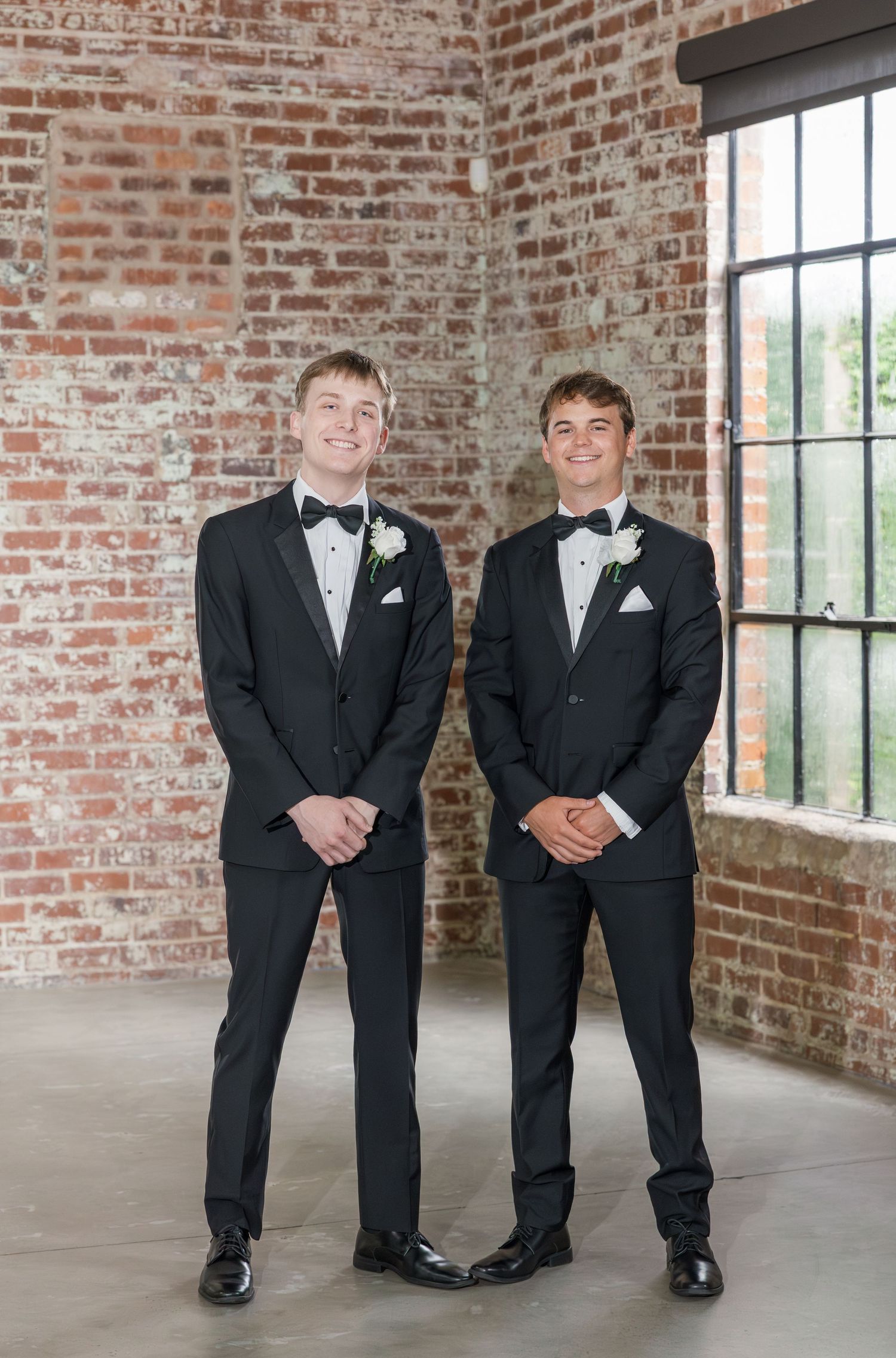 Two groomsmen in black tuxedos with boutonnieres pose against a rustic brick wall.