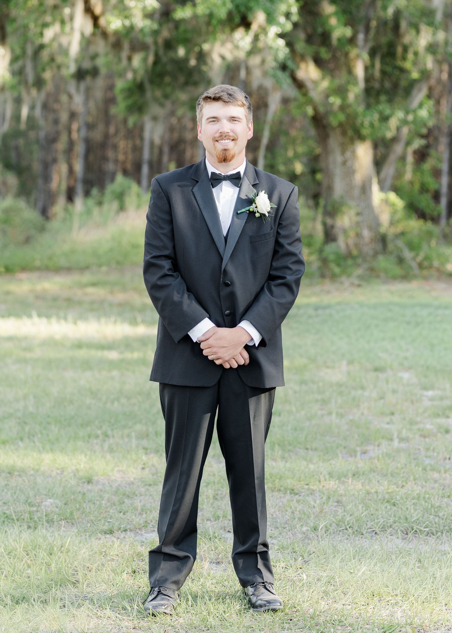 Groom in black tuxedo with white boutonniere standing on grass with trees in background.