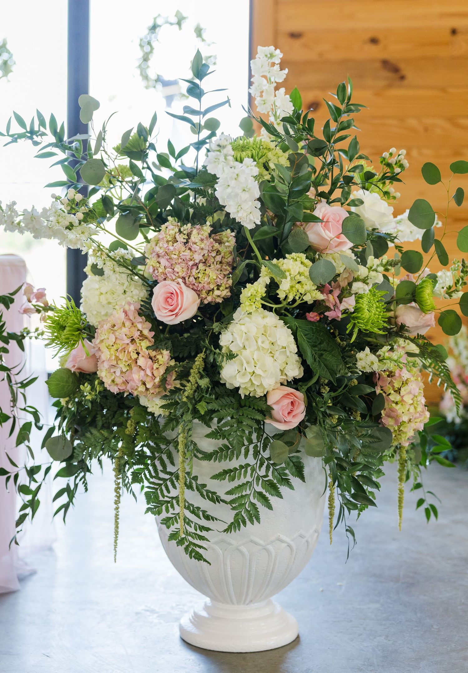 Lush wedding floral arrangement with pink roses, white hydrangeas, and greenery in white urn vase.