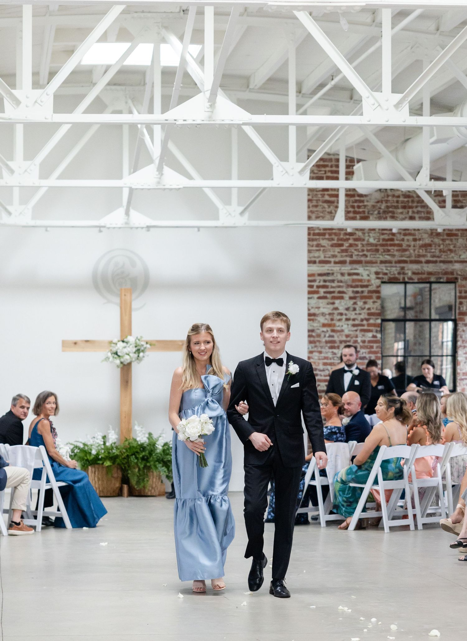 Groomsman escorts bridesmaid in blue gown down the aisle of an elegant industrial wedding venue.