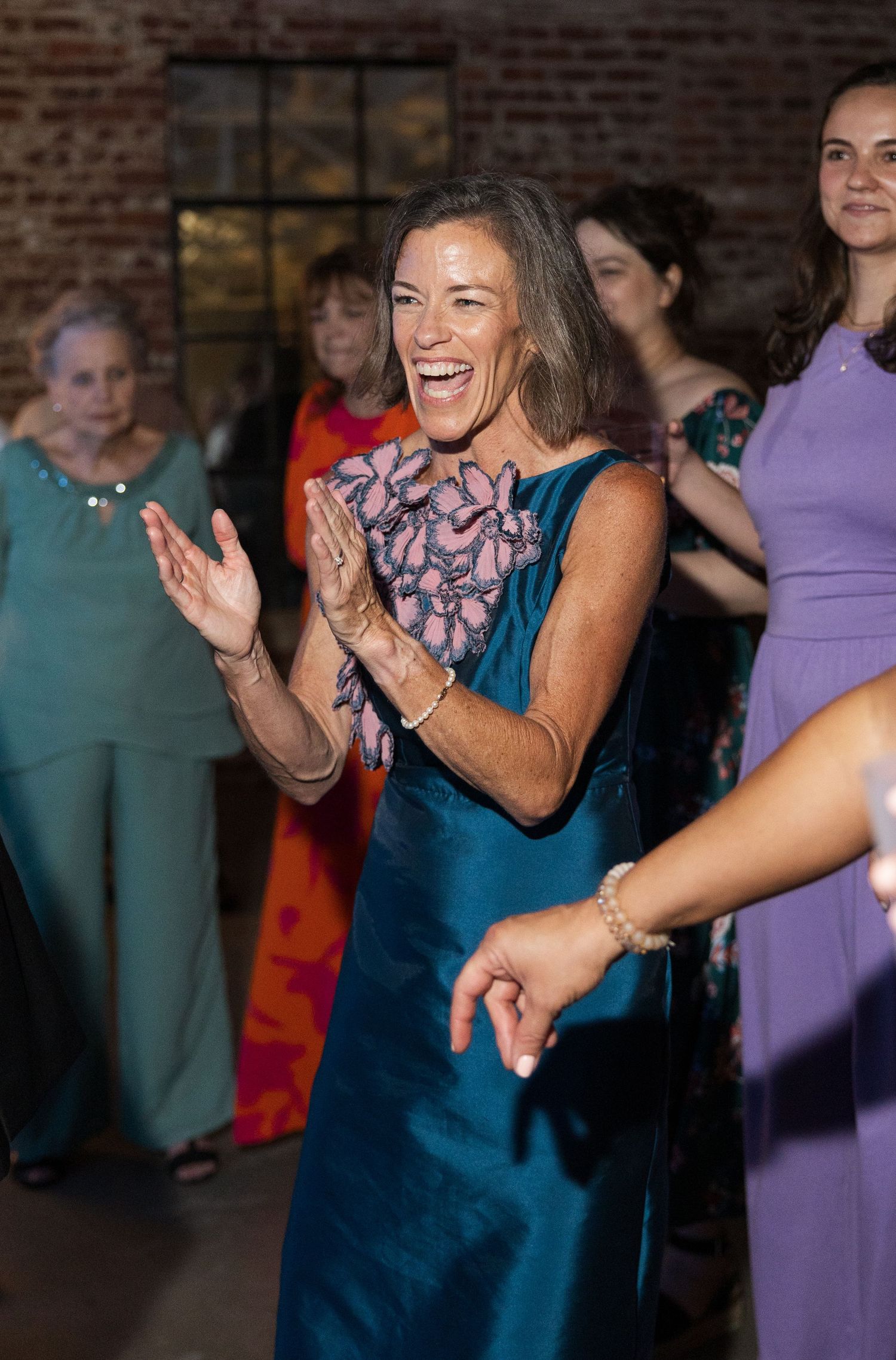 Joyful woman in teal floral dress claps and dances at elegant wedding reception celebration.