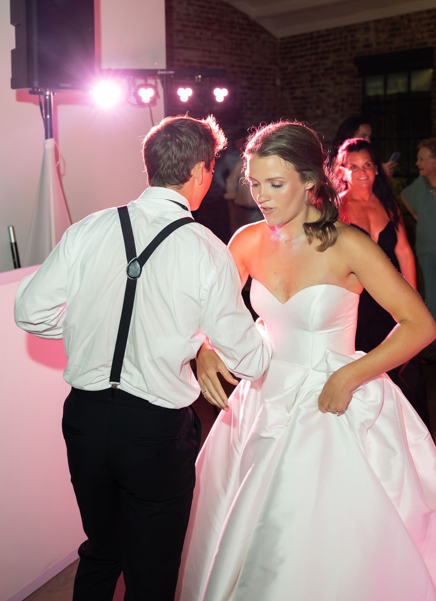 Bride in white strapless gown dances with groom in white shirt and suspenders at wedding reception.