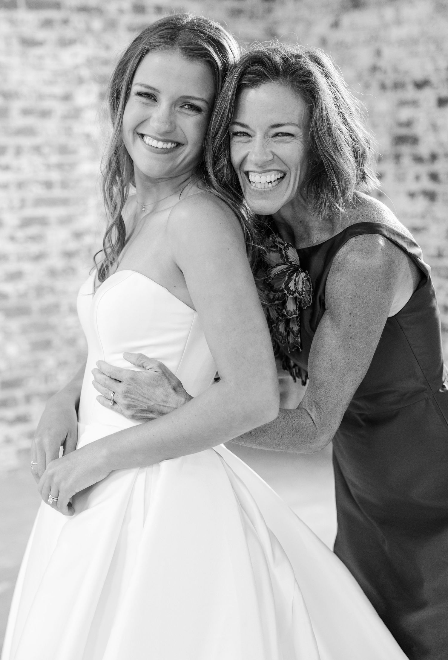 Black and white photo of smiling bride in strapless gown embracing a laughing woman against brick wall.