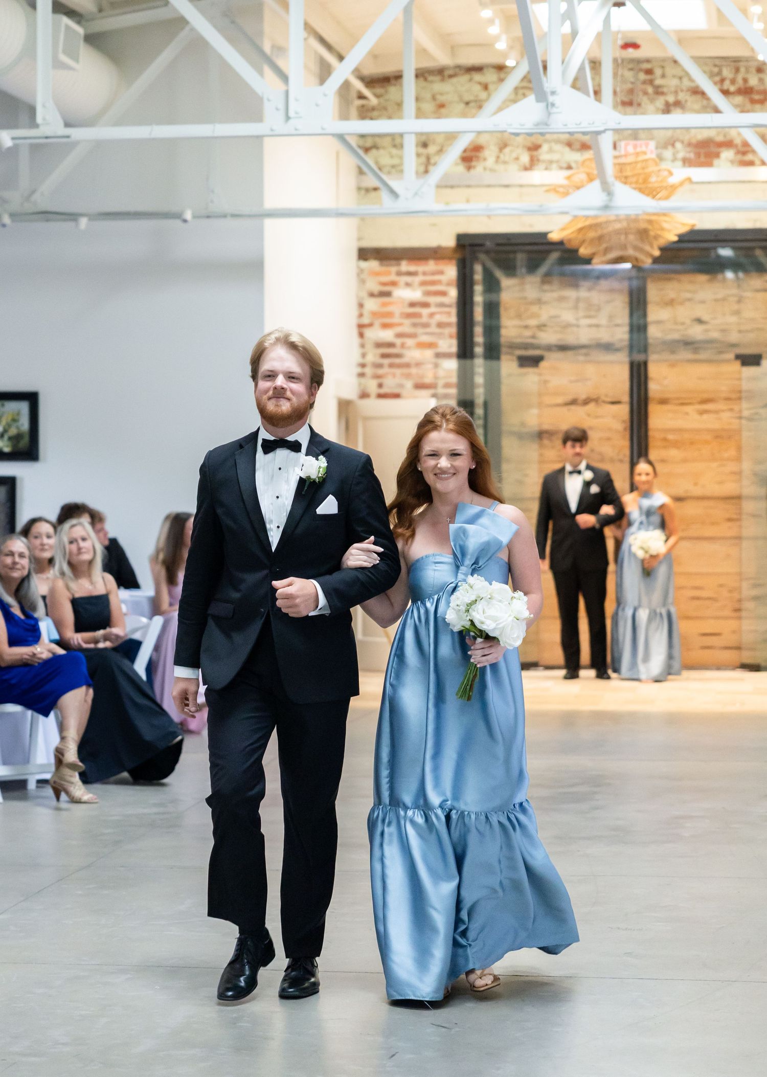 Groomsman in black tuxedo escorts bridesmaid in blue gown down wedding ceremony aisle.