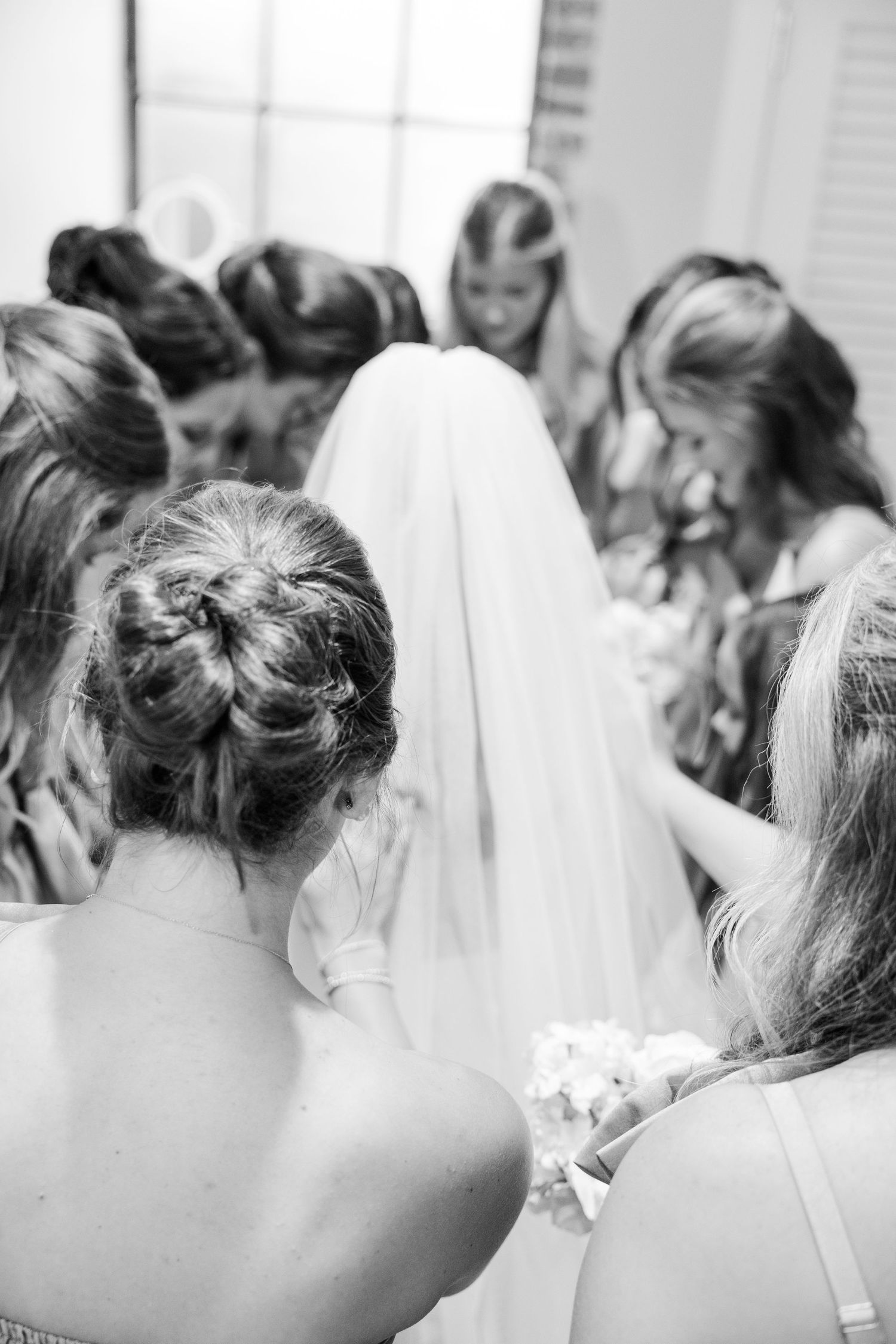 Black and white photo of bridesmaids surrounding bride adjusting her veil from behind.