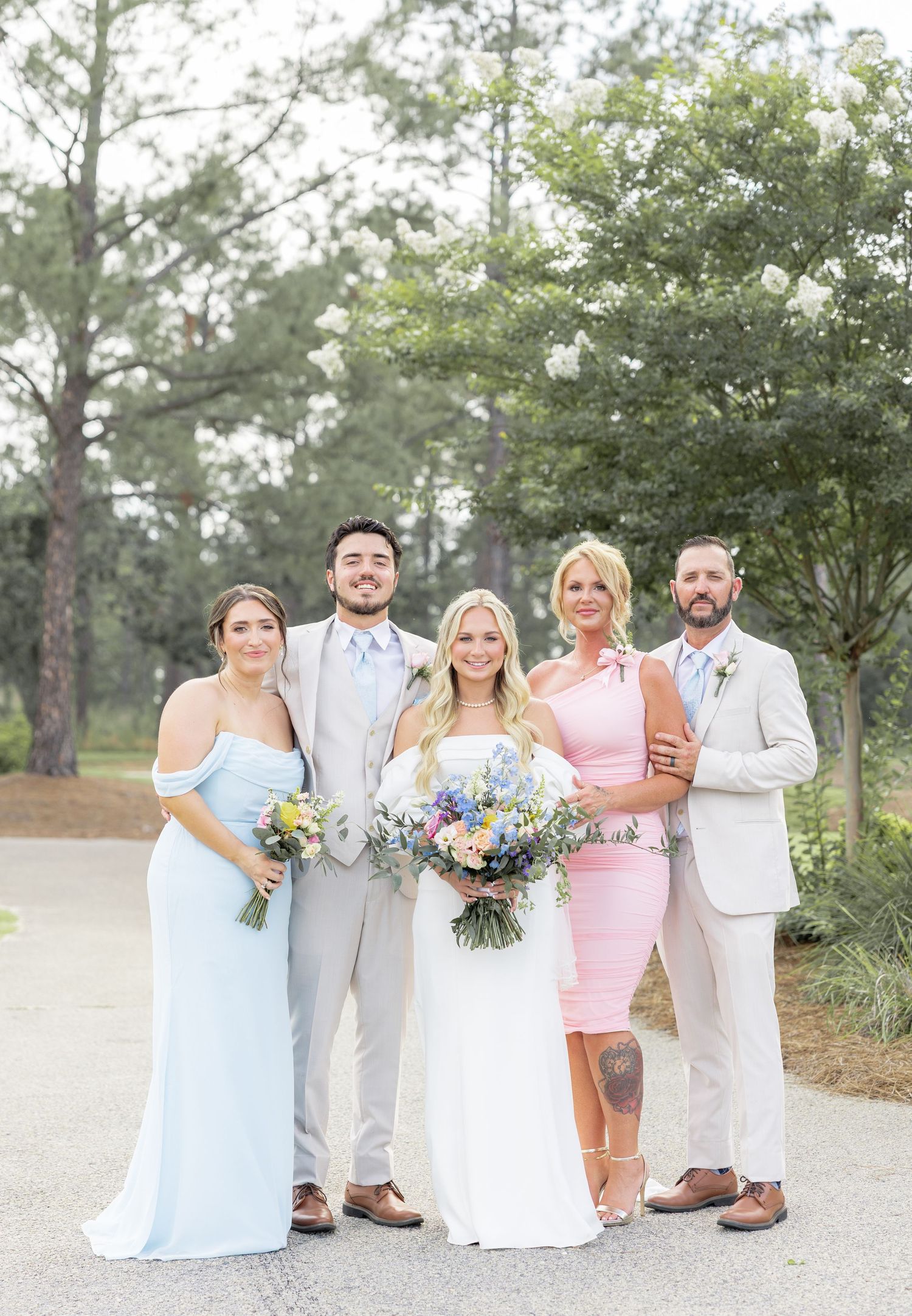 A wedding party of five poses together outdoors, featuring the bride in white and guests in blue and pink.